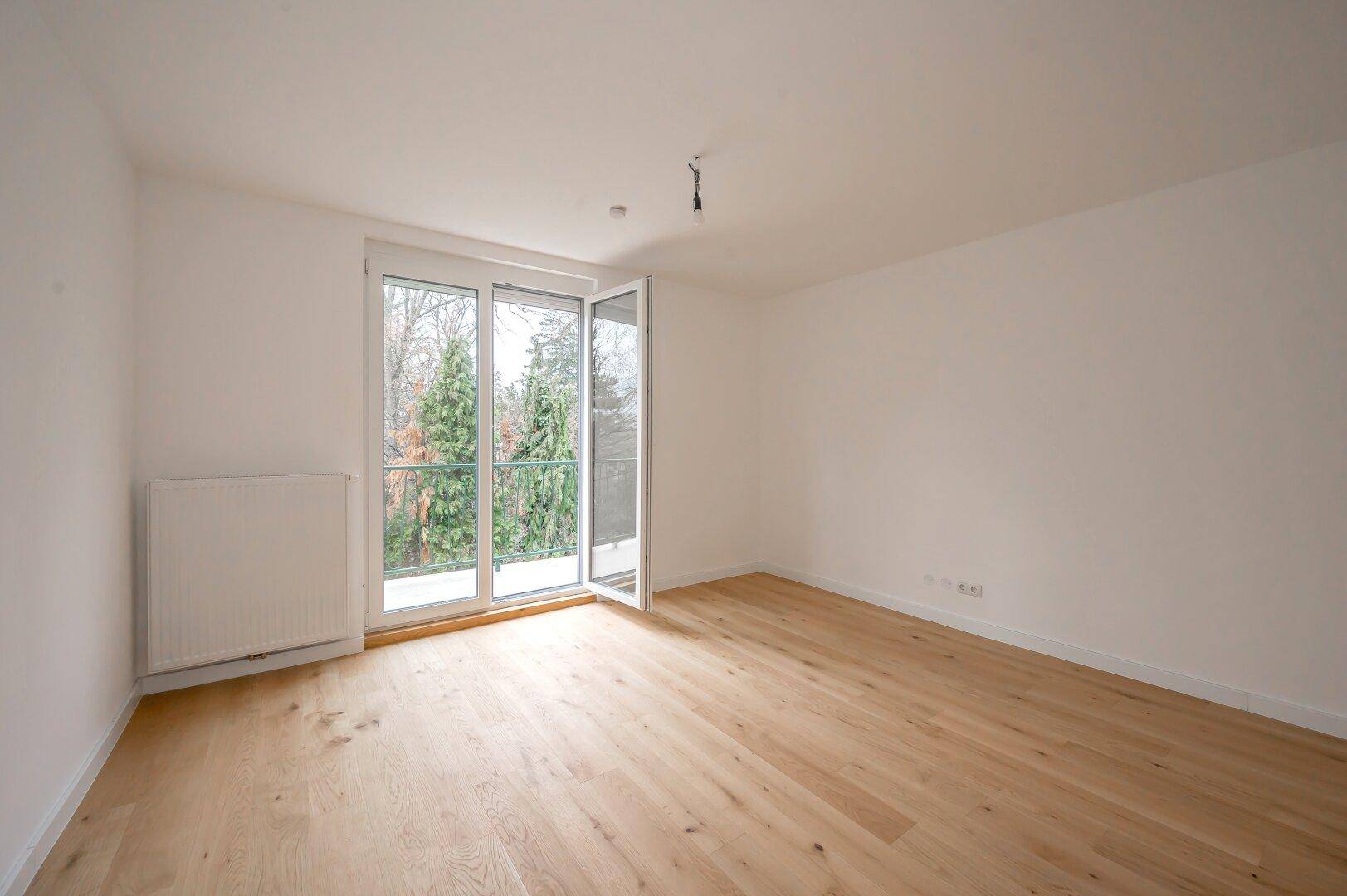 Leeres Zimmer mit hellem Holzboden, großen Fenstern und Blick ins Grüne.