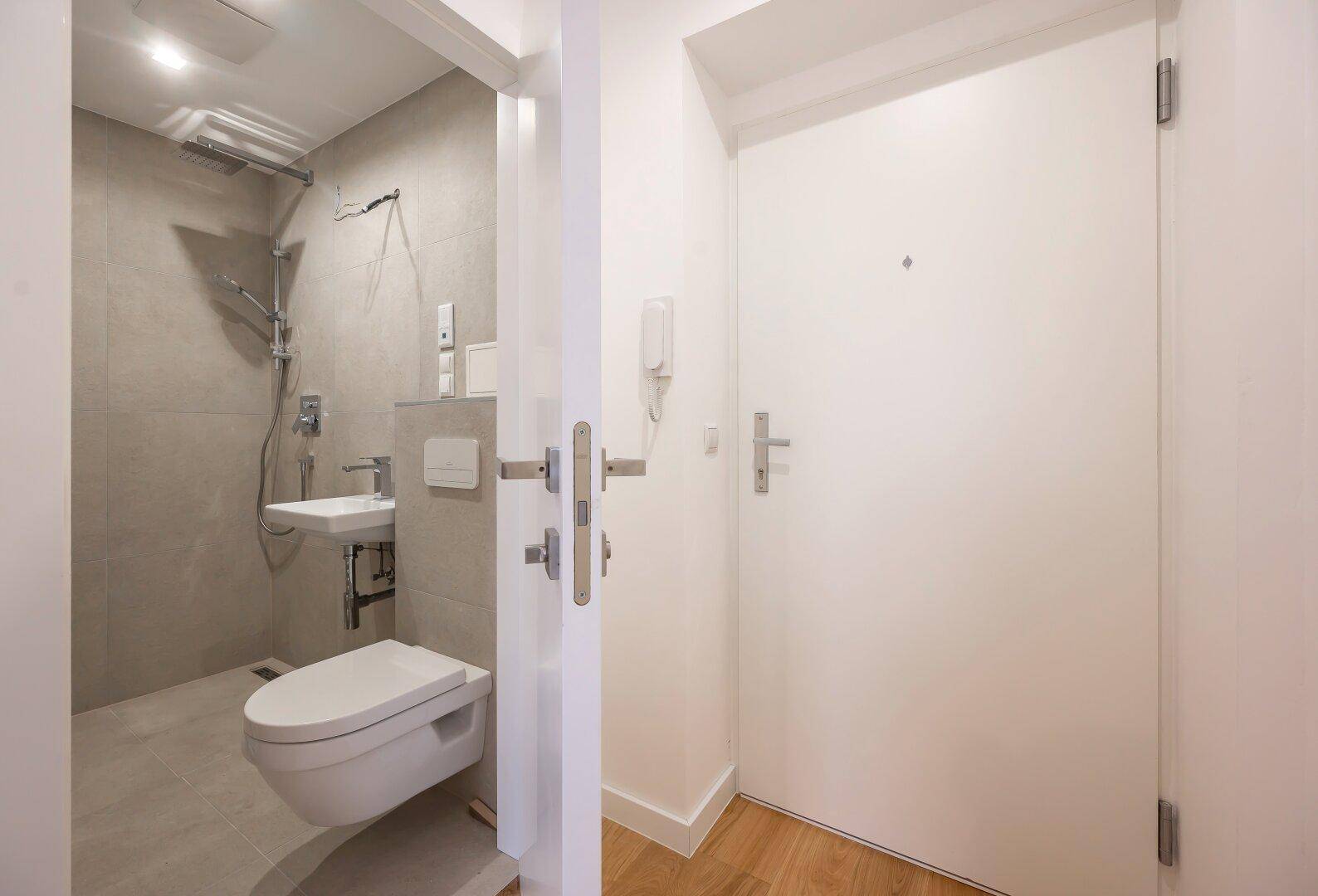 Kompaktes Badezimmer mit wandmontierter Toilette, kleinem Waschbecken und Dusche.