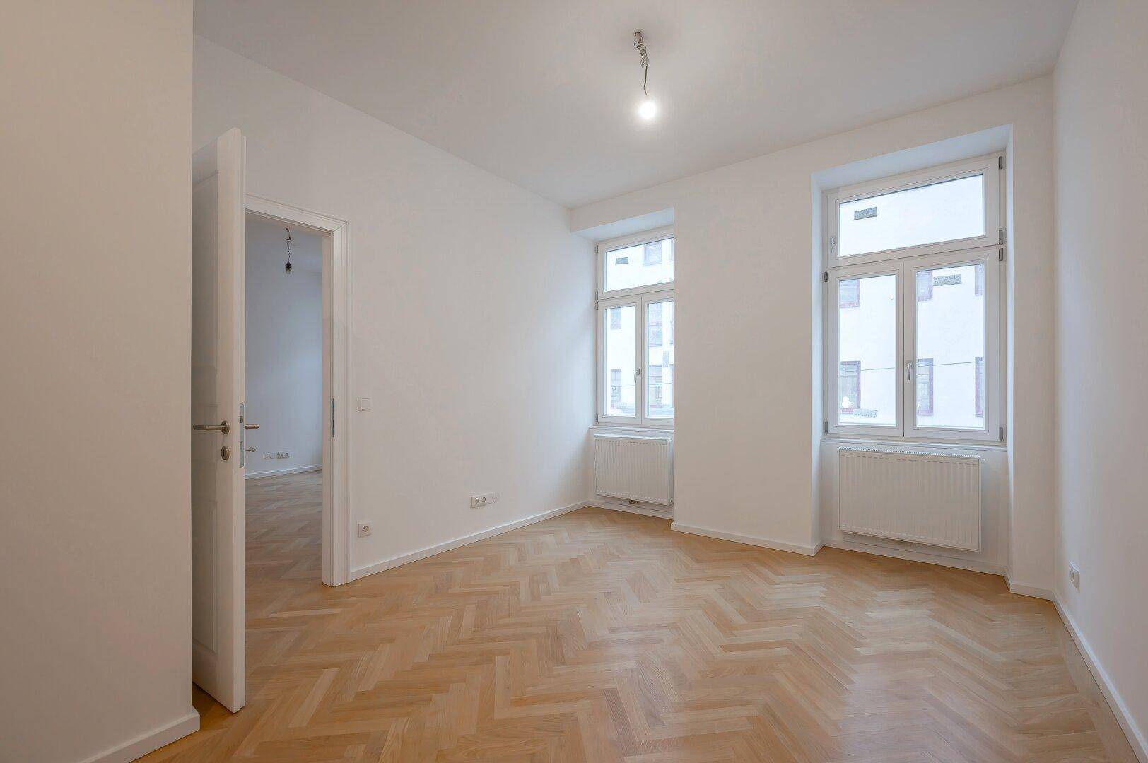 Unmöbliertes Schlafzimmer mit Fischgrätparkett, hohen Decken und zwei Fenstern.
