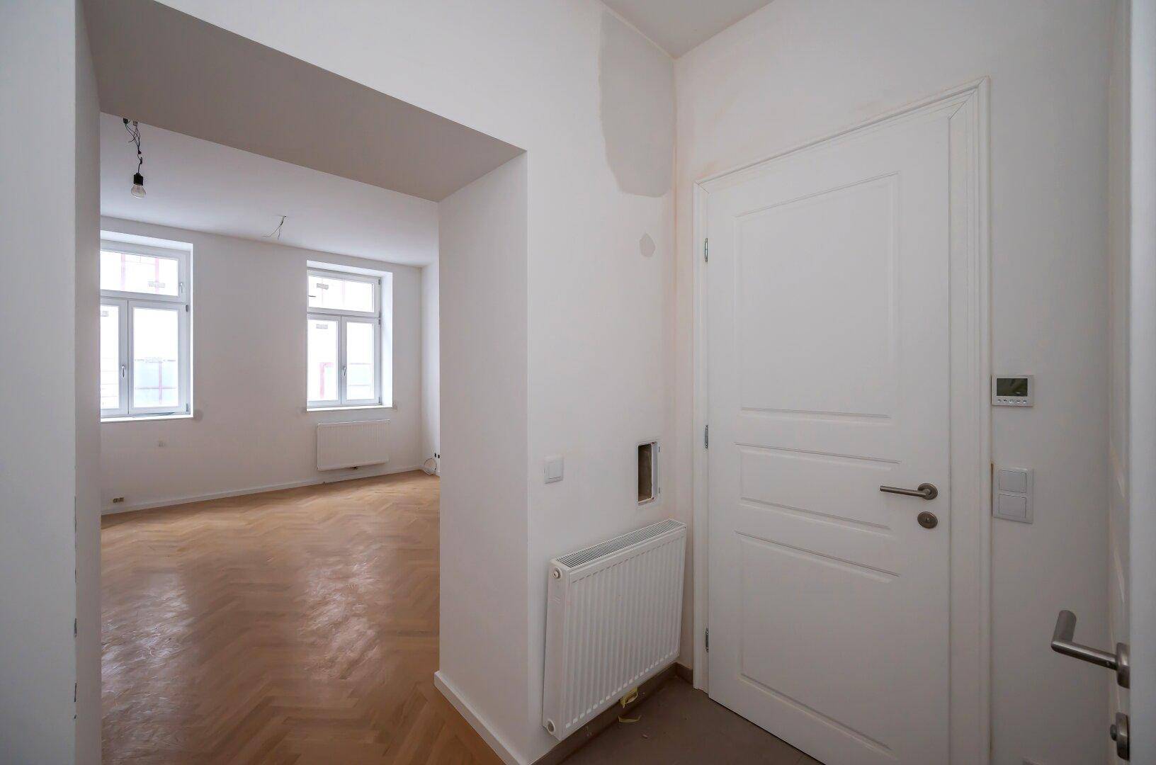 Heller Flur mit weißer Tür, Heizkörper und Blick in ein angrenzendes Zimmer mit Parkettboden.