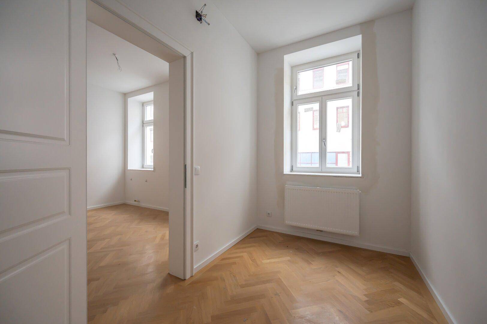 Ruhiges Zimmer mit Fischgrätparkett, weiß gestrichenen Wänden und einem Fenster, ideal als Schlafzimmer.