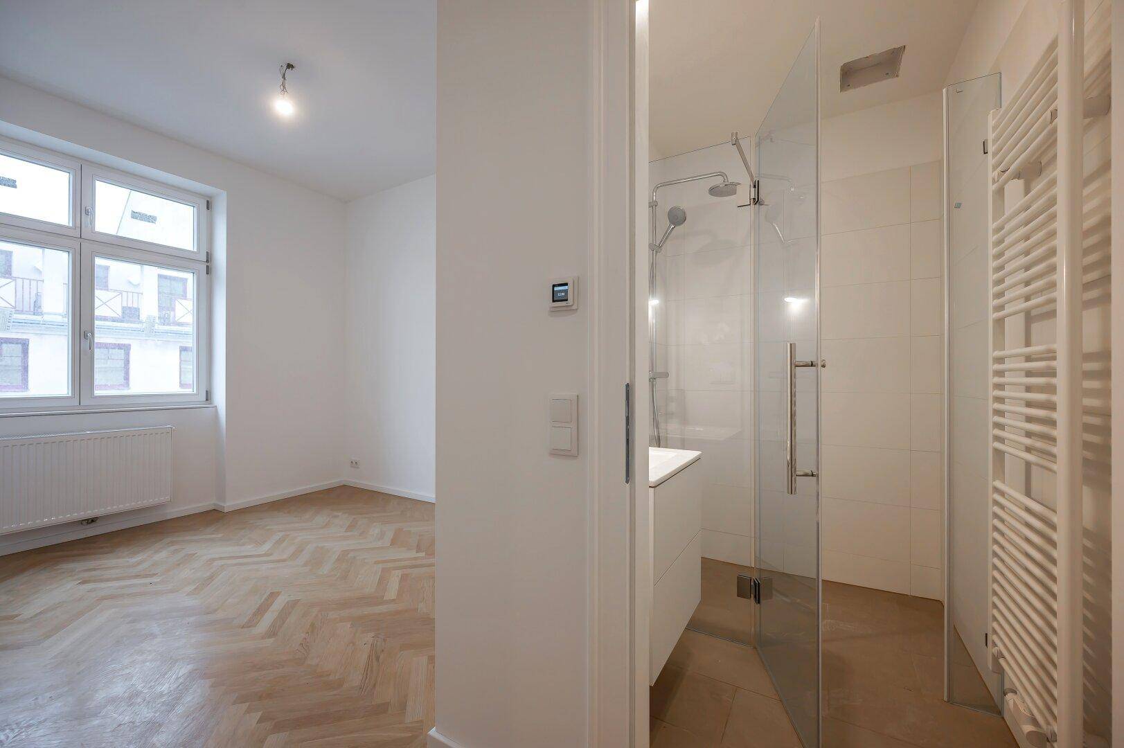 Modernes Badezimmer mit begehbarer Dusche und Zugang zu einem hellen Zimmer mit Parkett.