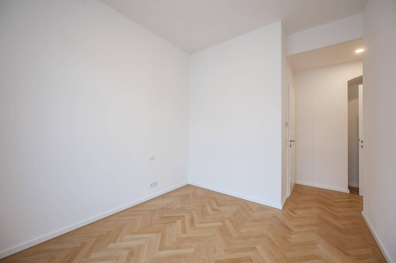 Leerer Raum mit Fischgrätparkett und Übergang zu einem weiteren Bereich der Wohnung.