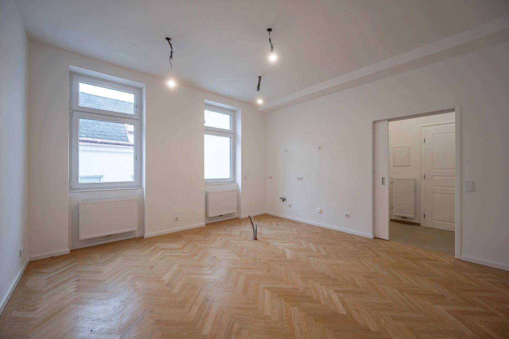 Geräumiges Wohnzimmer mit Fischgrätparkett, hohen Decken und zwei großen Fenstern.