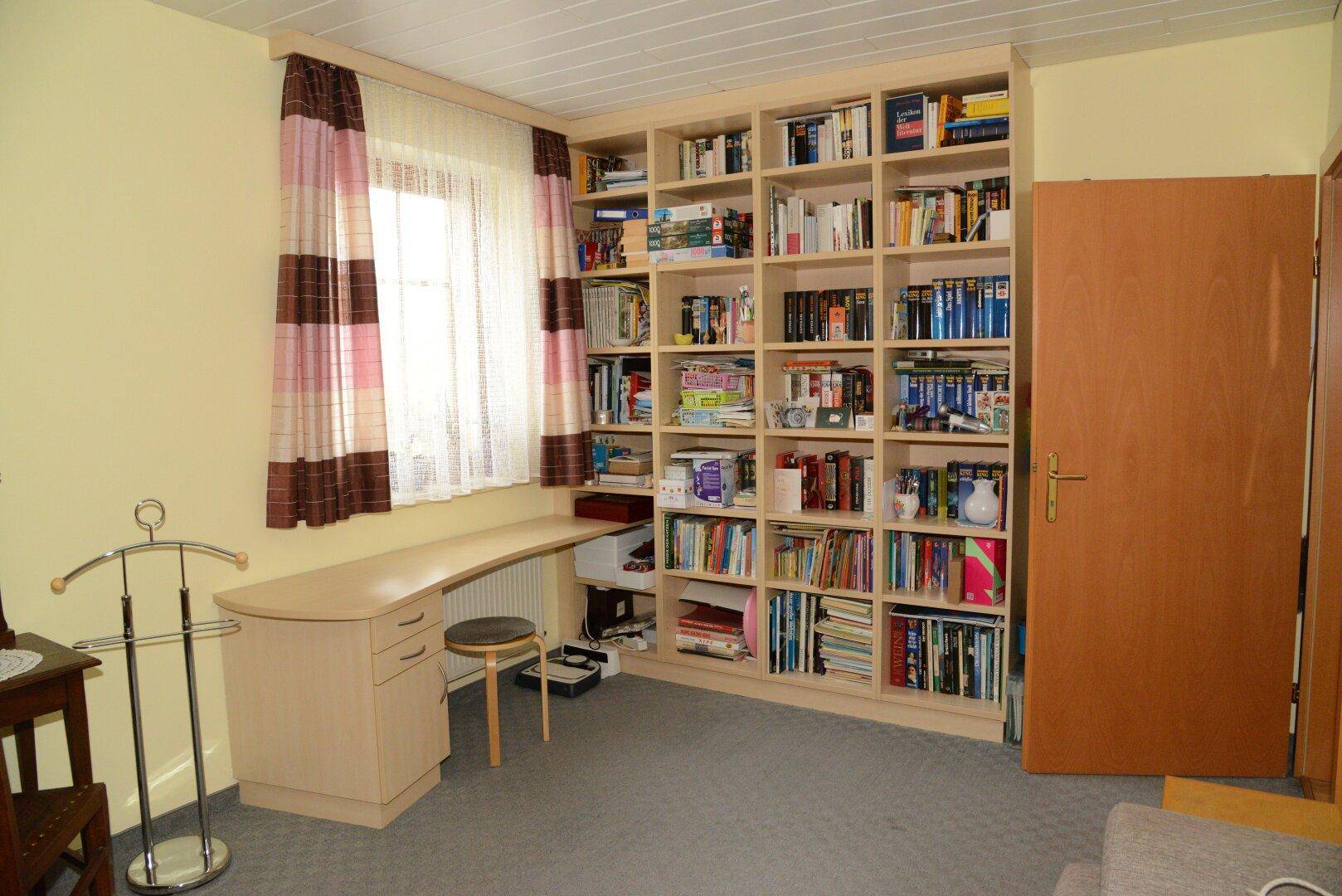 Arbeitszimmer mit großem Bücherregal, Schreibtisch und Fenster.