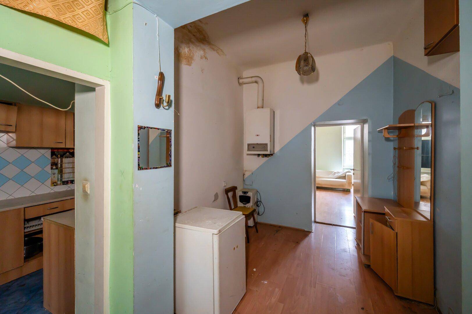 Flur mit Gastherme, Kühlschrank und Zugang zu Küche und weiteren Zimmern, renovierungsbedürftig.