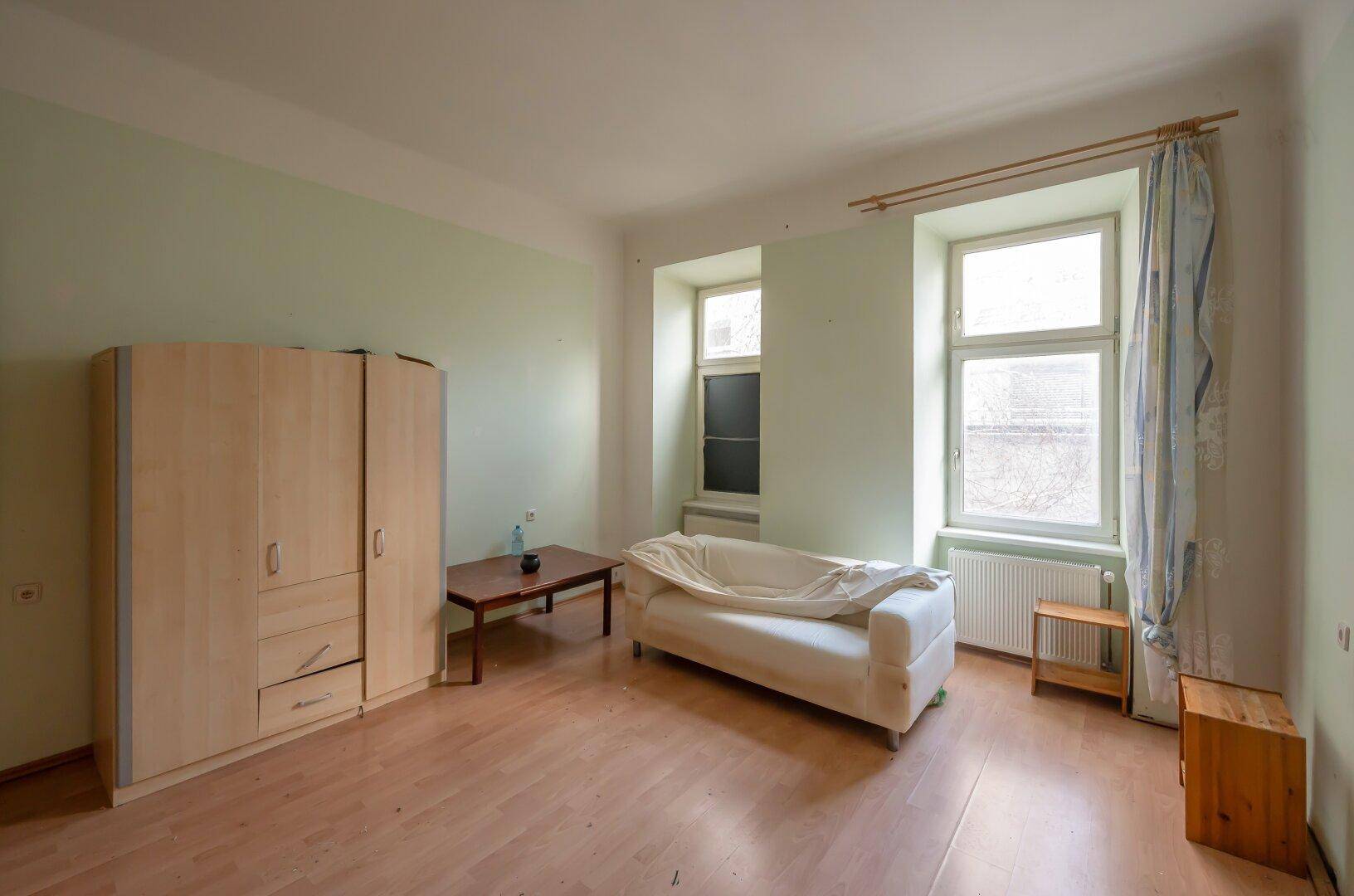 Geräumiges Wohnzimmer mit hellgrünen Wänden, Laminatboden und einfacher Einrichtung, bereit für eine Renovierung.