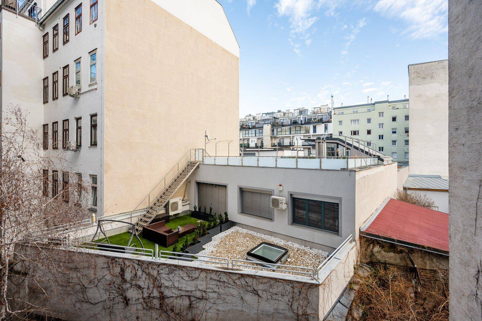 Dachterrasse mit Grünfläche, Kiesbereich und Schaukel, umgeben von Gebäuden.