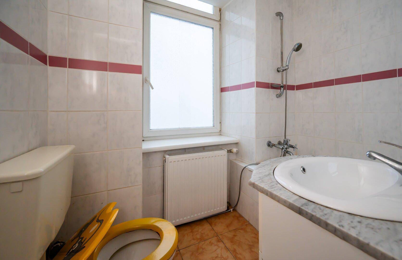 Badezimmer mit WC, Waschbecken, Duschvorrichtung und Fenster, gefliest.
