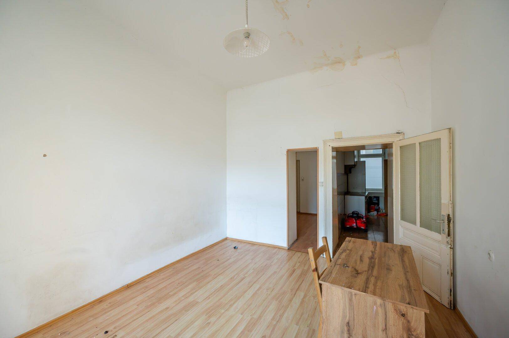 Einfaches Zimmer mit Laminatboden und Zugang zu einem weiteren Raum, Decke mit Flecken.
