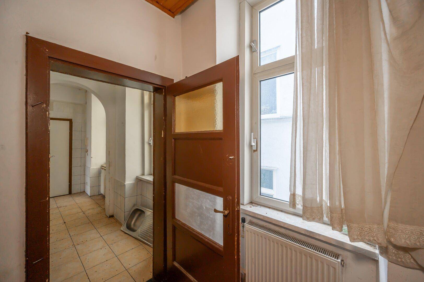 Flur mit gefliestem Boden, Fenster und einer alten Holztür, der Renovierungsbedarf aufweist.