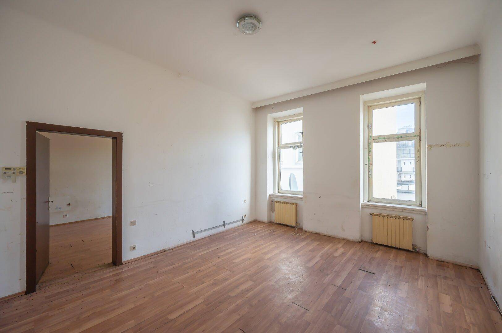 Leeres Zimmer mit Holzboden und zwei Fenstern, das Renovierungsbedarf aufweist.