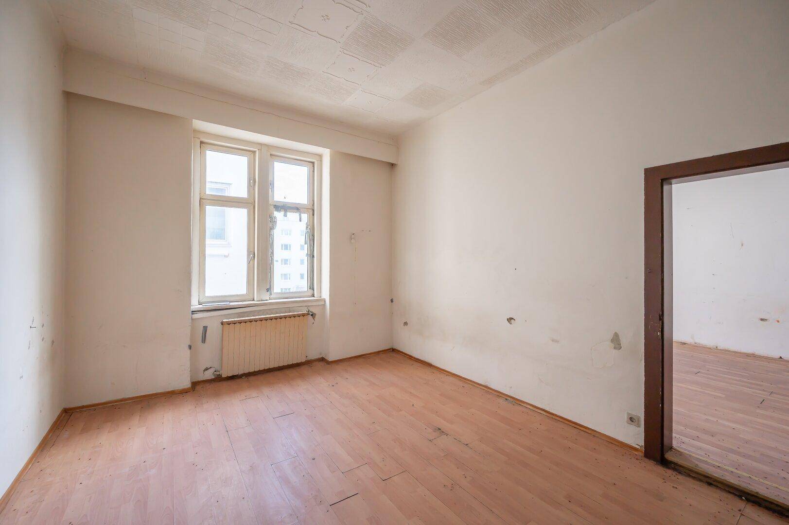 Leeres Zimmer mit Holzboden und einem Fenster, das deutlichen Renovierungsbedarf zeigt.