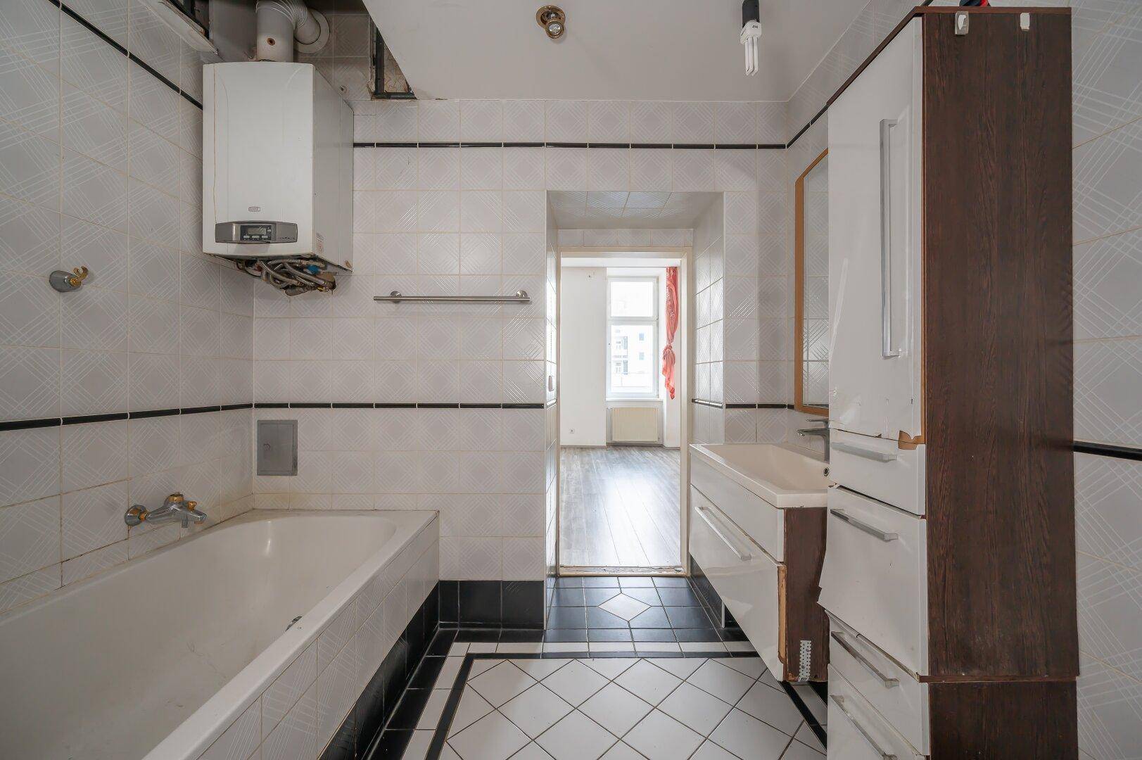 Badezimmer mit Badewanne, Waschbecken und Warmwasserbereiter, Blick in einen weiteren Raum.