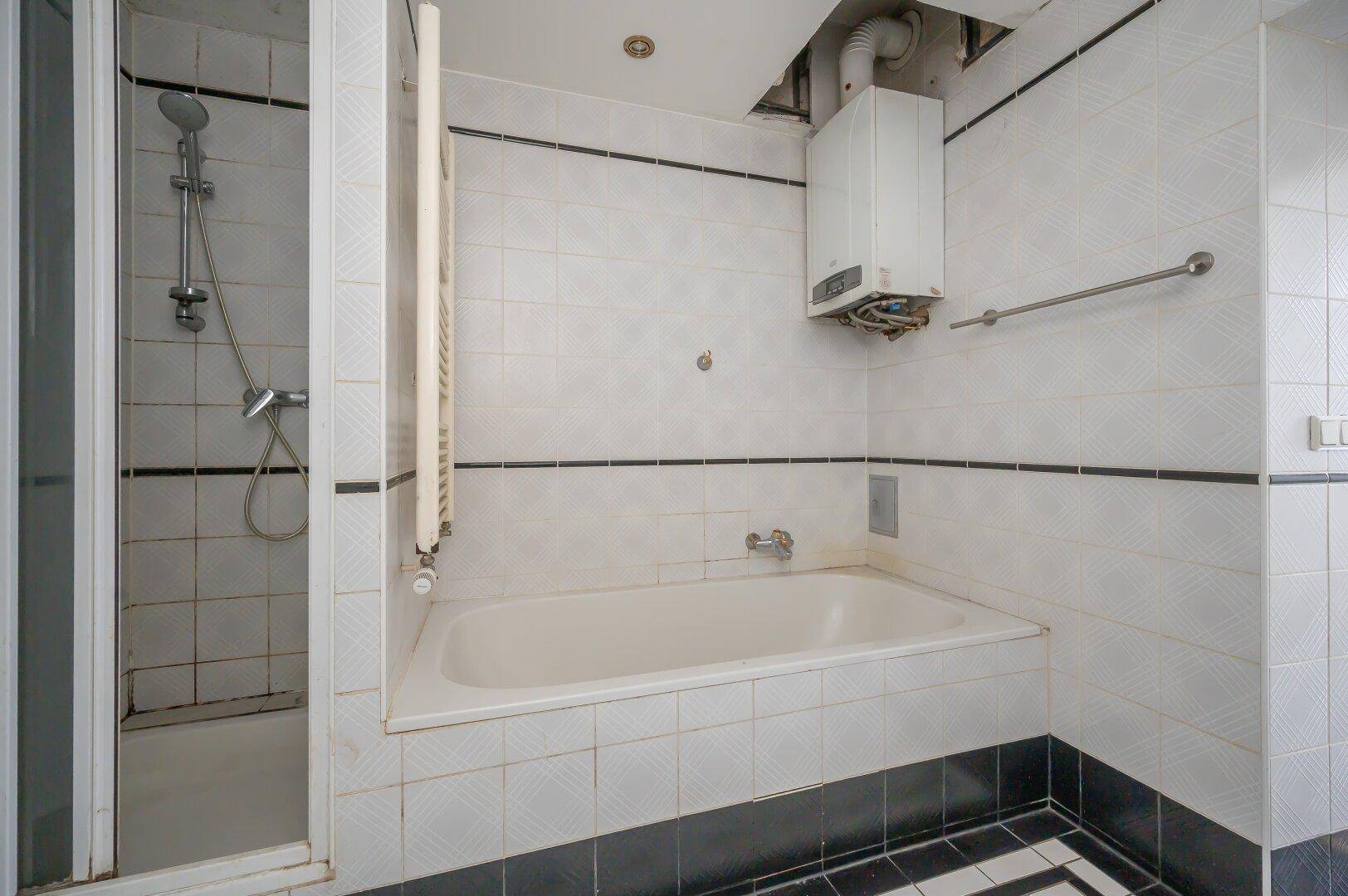 Badezimmer mit Badewanne und Duschkabine, zeigt ältere Fliesen und einen Warmwasserbereiter.