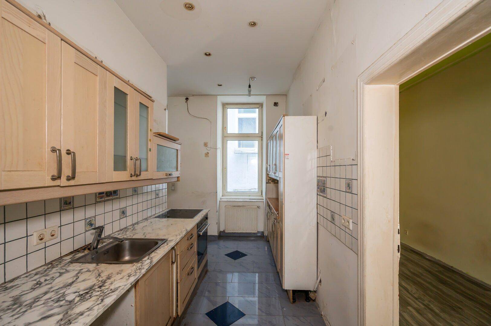 Schmale Küche mit älteren Einbauschränken, Fliesenboden und einem Fenster.
