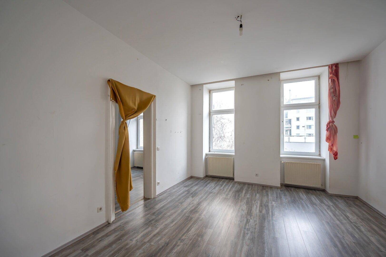 Unmöbliertes Zimmer mit Laminatboden und zwei Fenstern, bereit für eine Renovierung.