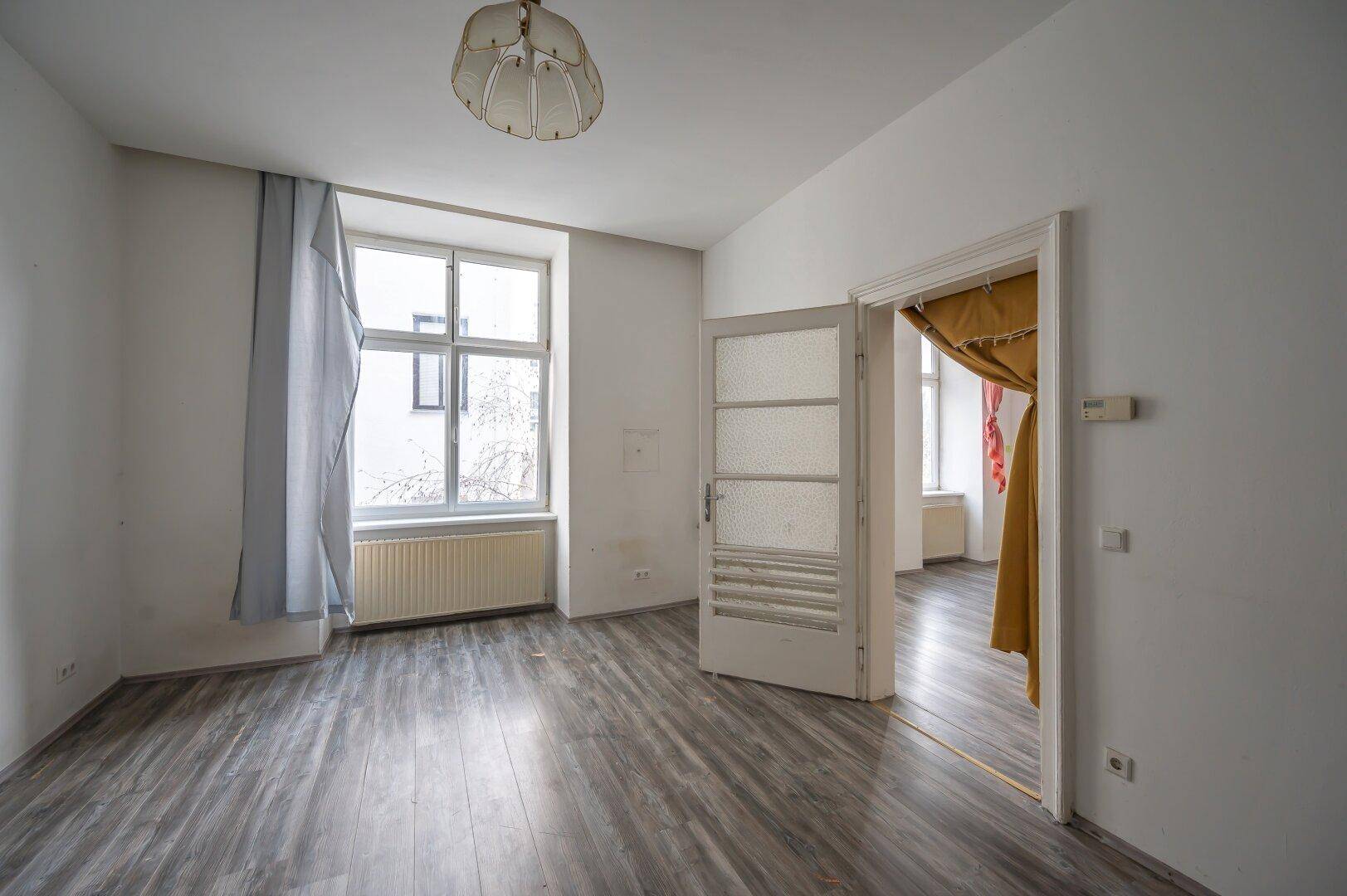 Leeres Wohnzimmer mit Laminatboden, großem Fenster und einer Tür zu einem weiteren Raum.