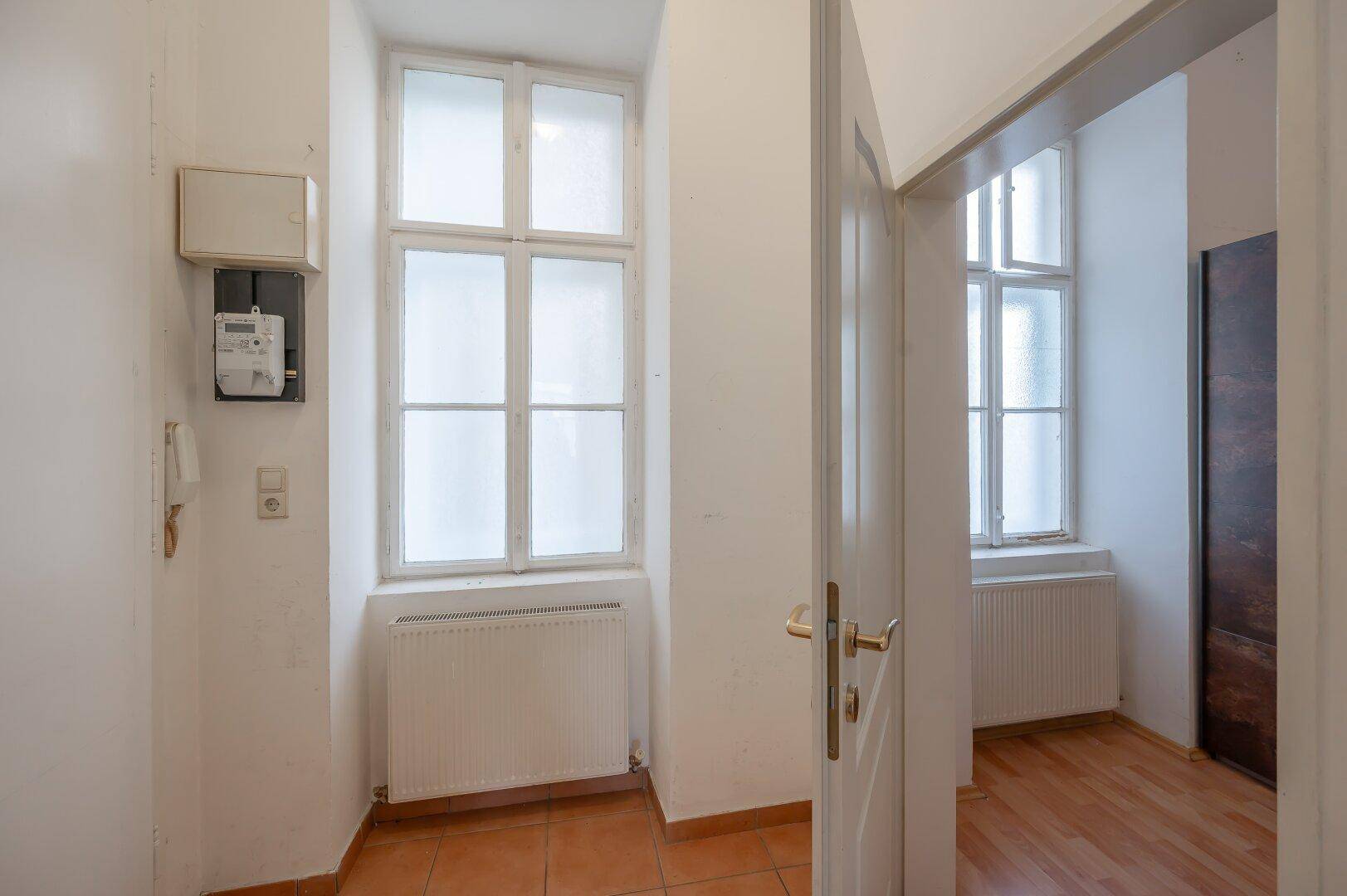 Eingangsbereich mit Fliesenboden, Fenstern und einem Zählerkasten an der Wand.
