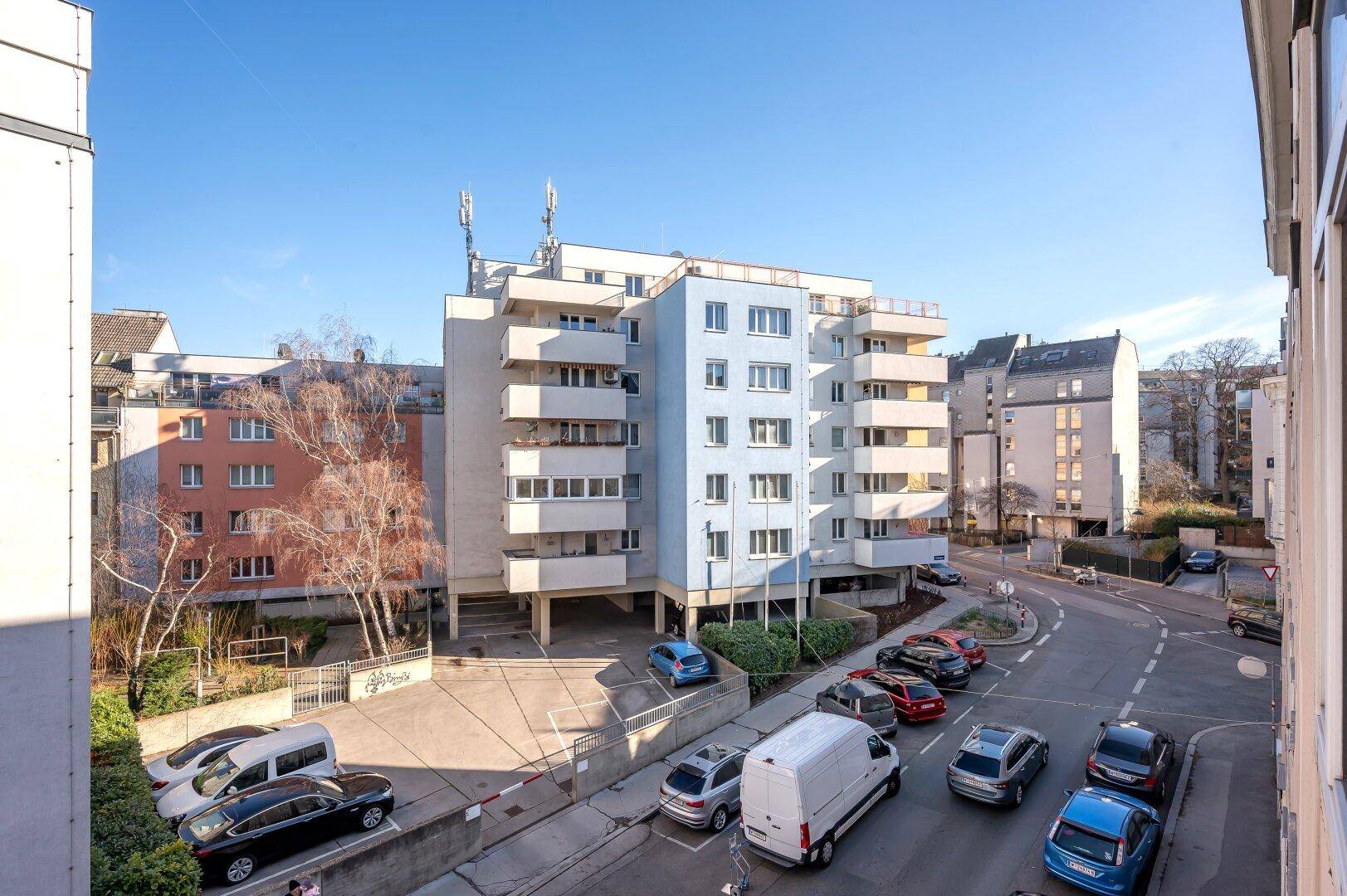 Mehrfamilienhaus mit Balkonen und Parkplätzen an einer städtischen Straße unter blauem Himmel.