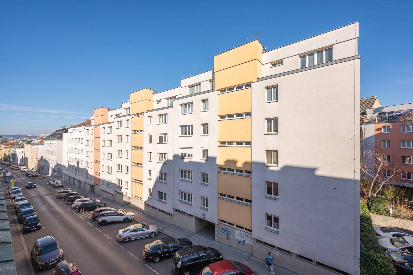 Lange Wohngebäudezeile mit Balkonen und Parkplätzen entlang einer städtischen Straße.