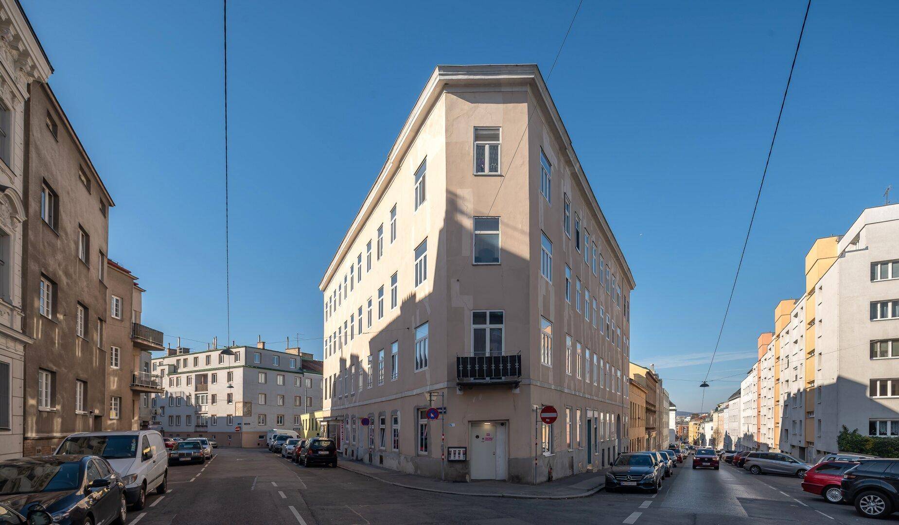 Eckgebäude in einer städtischen Umgebung mit mehreren Stockwerken und Fenstern, unter klarem Himmel.