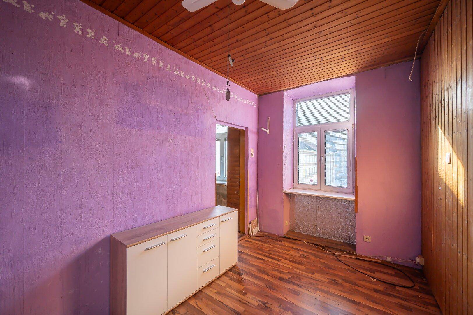 Schlafzimmer mit lila Wänden, Holzbalkendecke und einem Deckenventilator, bereit zur Modernisierung.