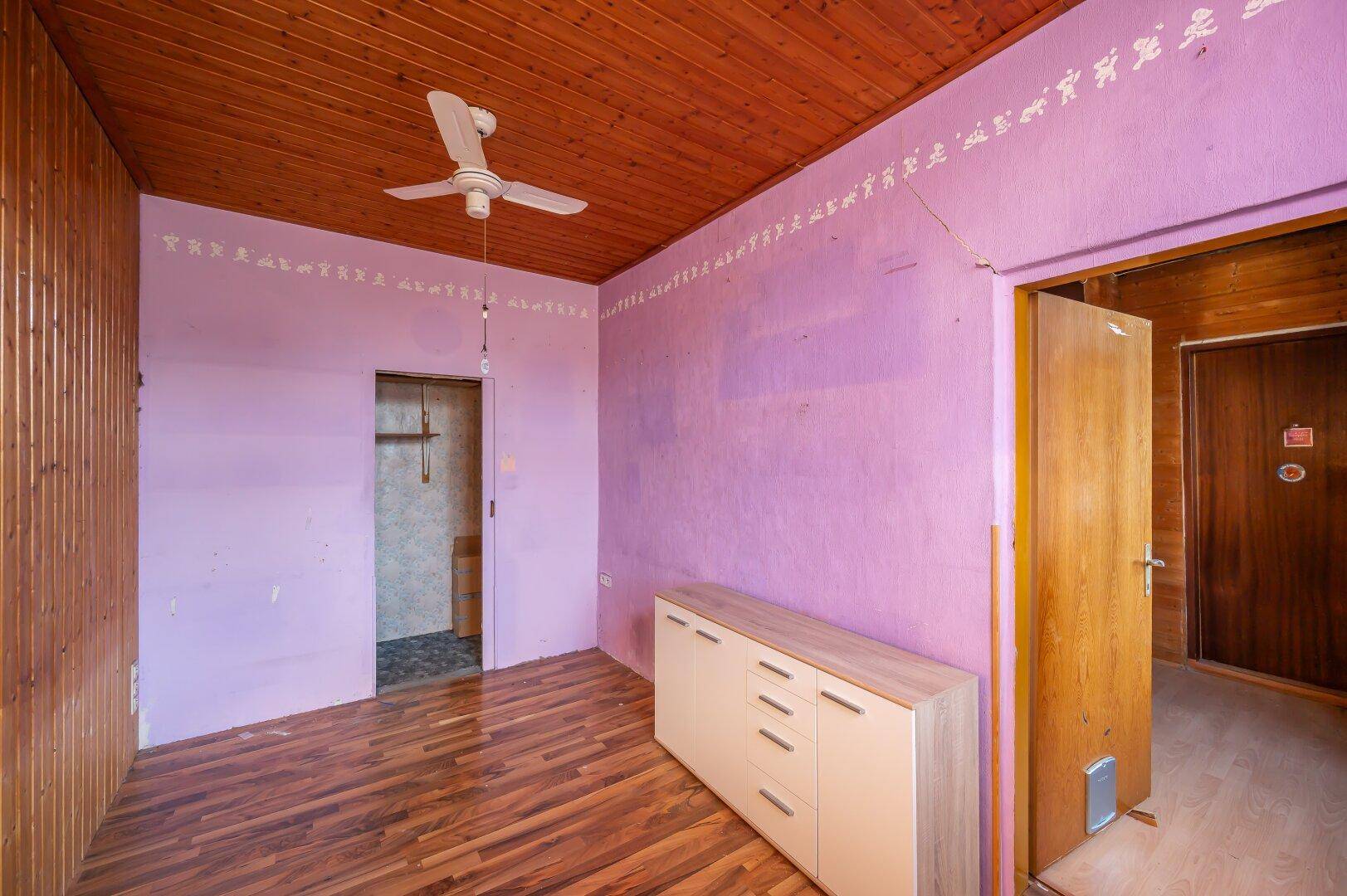 Schlafzimmer mit lila Wänden, Holzbalkendecke und Deckenventilator, bietet Potenzial für individuelle Gestaltung.
