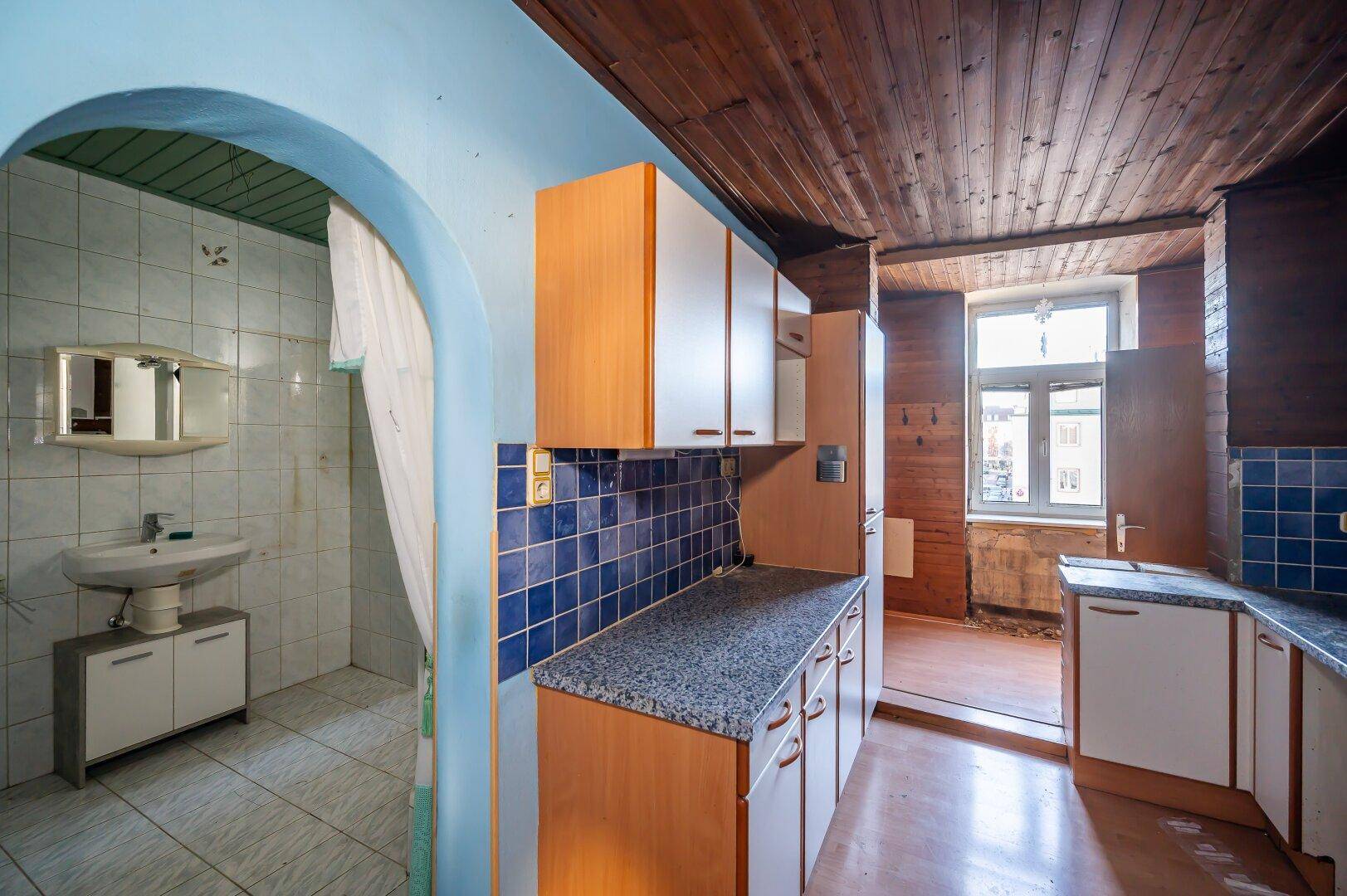 Küche mit blauen Fliesen, Holzschränken und angrenzendem Badezimmer, benötigt Modernisierung.