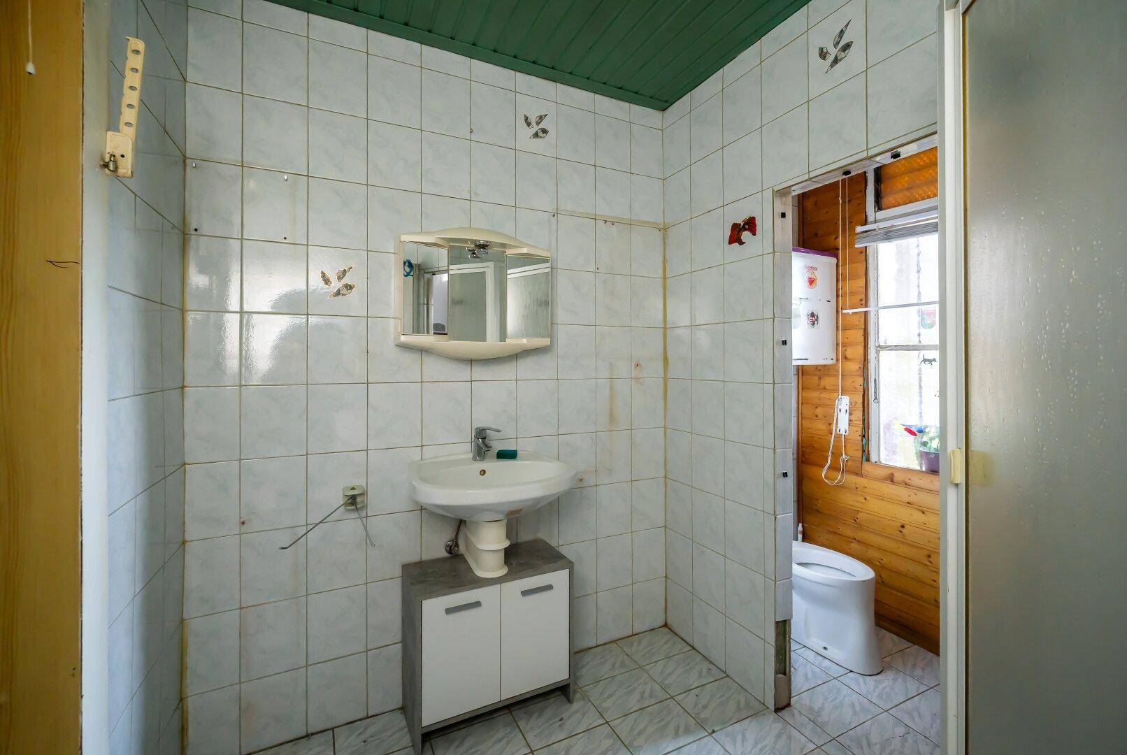 Gefliestes Badezimmer mit Waschbecken und Spiegel, angrenzend an einen Raum mit Toilette.