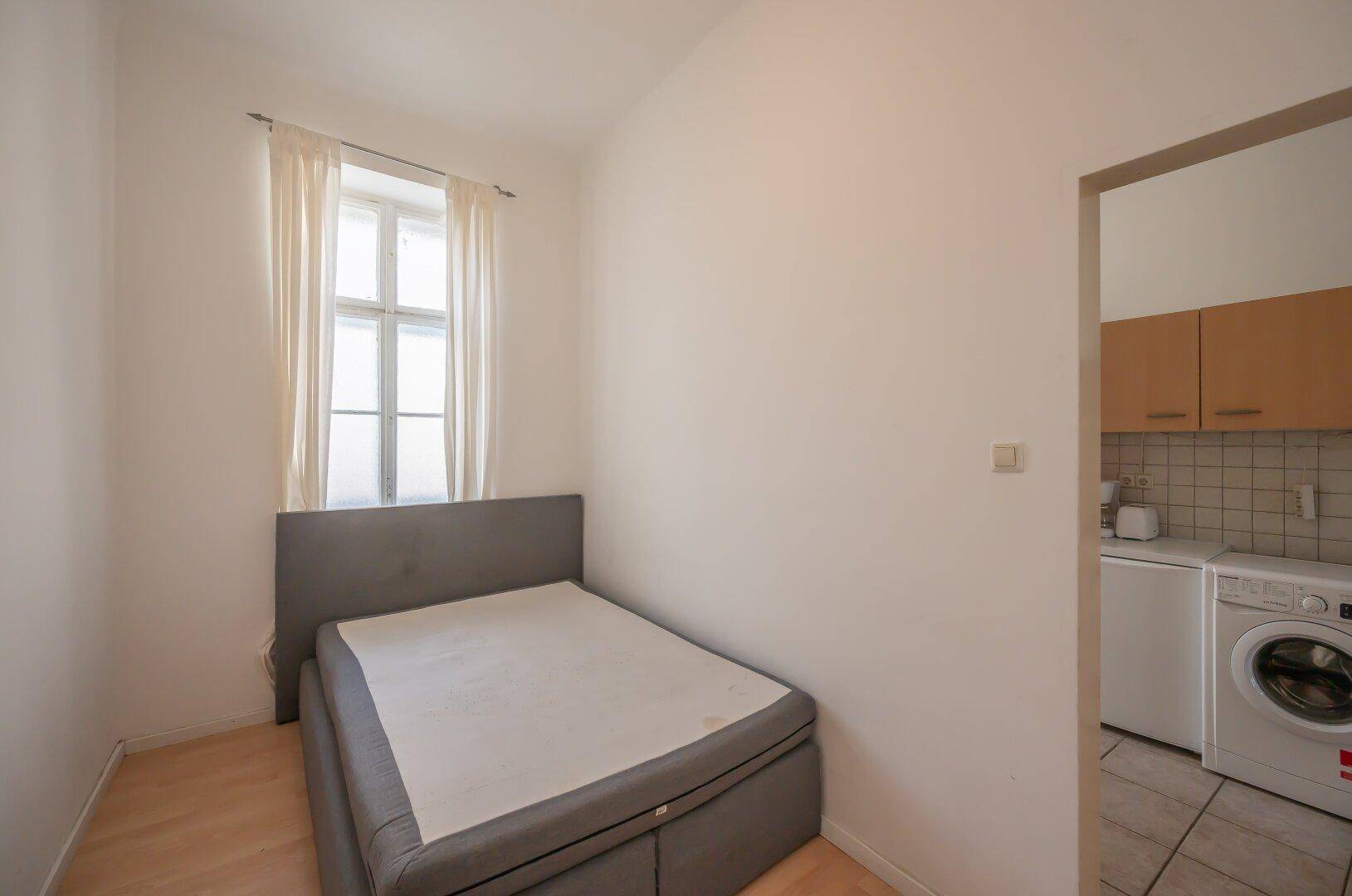 Kleines Schlafzimmer mit Einzelbett und Fenster, angrenzend an einen Bereich mit Waschmaschine.