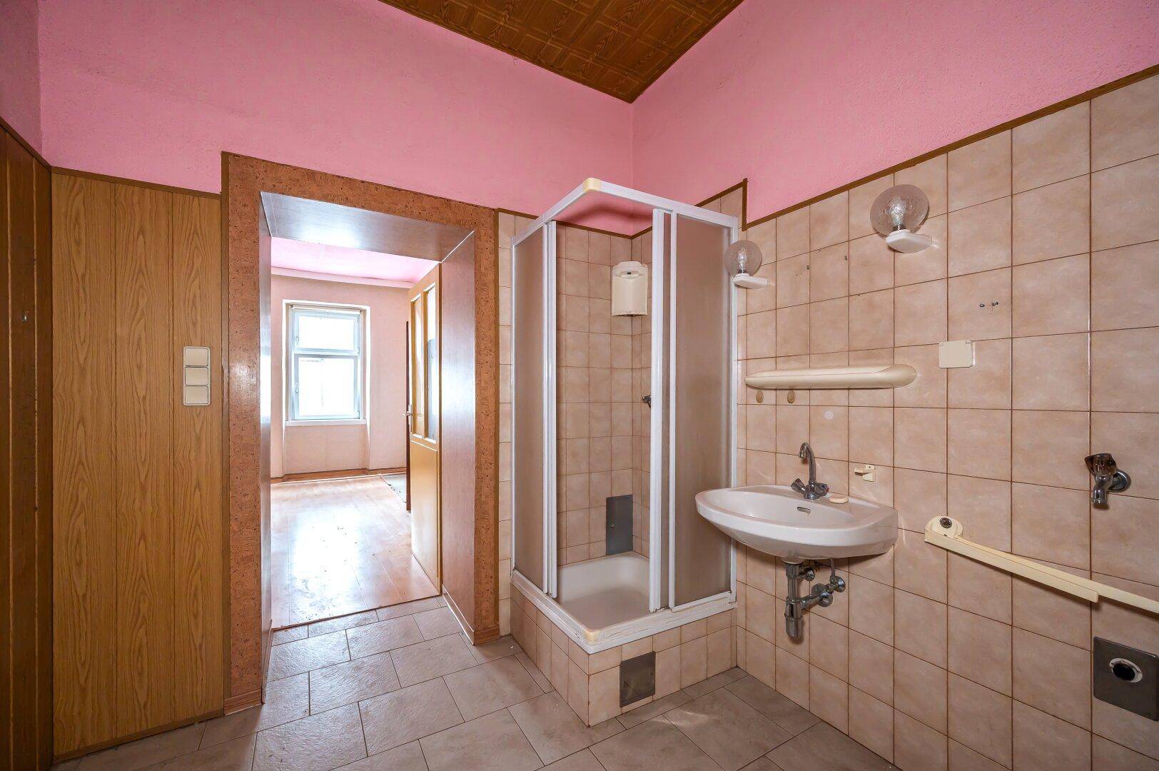 Veraltetes Badezimmer mit Duschkabine, Waschbecken und beigen Fliesen, renovierungsbedürftig.