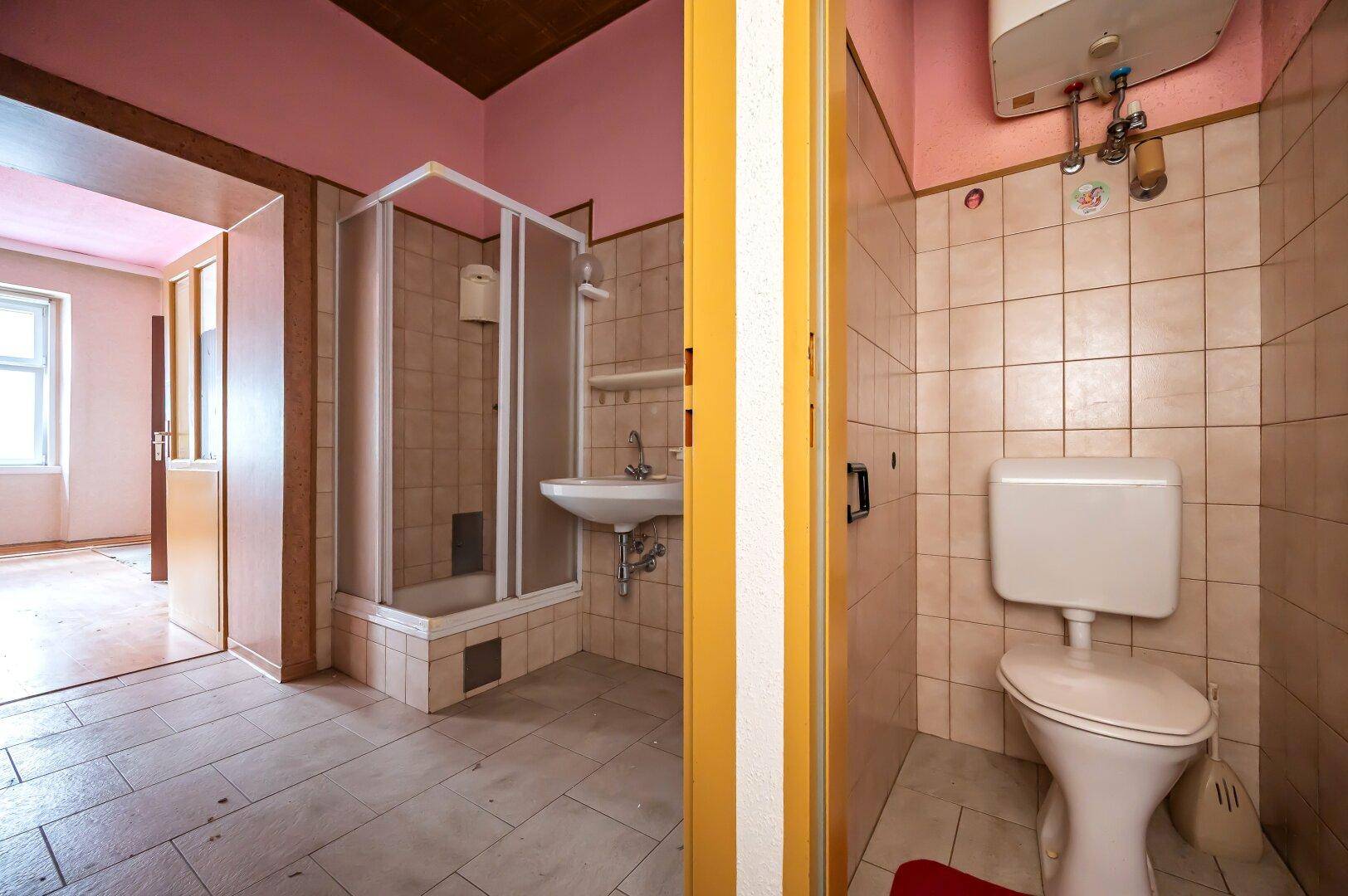 Veraltetes Badezimmer mit Duschkabine, Toilette, Waschbecken und beigen Fliesen, renovierungsbedürftig.