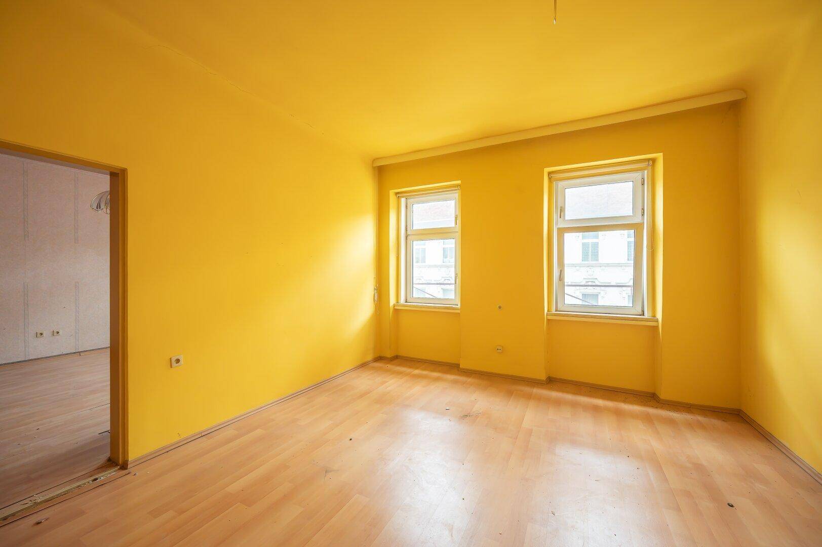 Helles Zimmer mit gelben Wänden, Laminatboden und zwei Fenstern, renovierungsbedürftig.