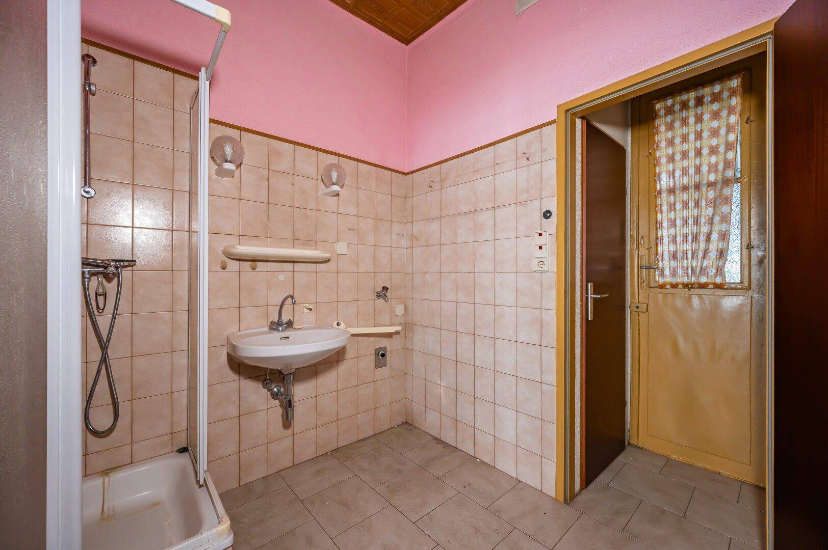 Veraltetes Badezimmer mit Duschkabine, Waschbecken und beigen Fliesen, renovierungsbedürftig.