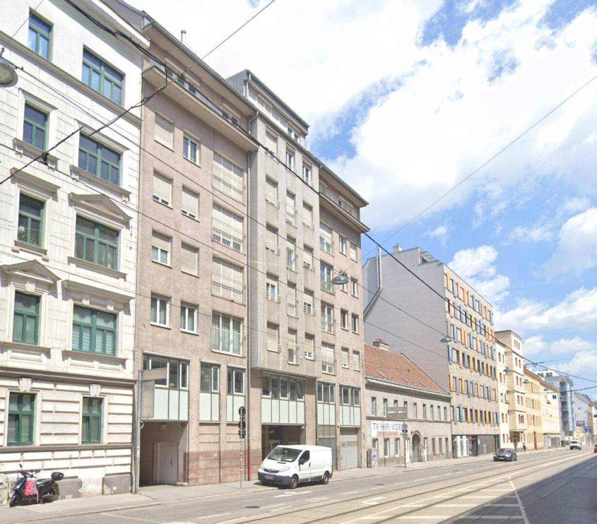 Eine belebte Stadtstraße mit verschiedenen Gebäuden, Fahrzeugen und einer Straßenbahnlinie unter blauem Himmel.