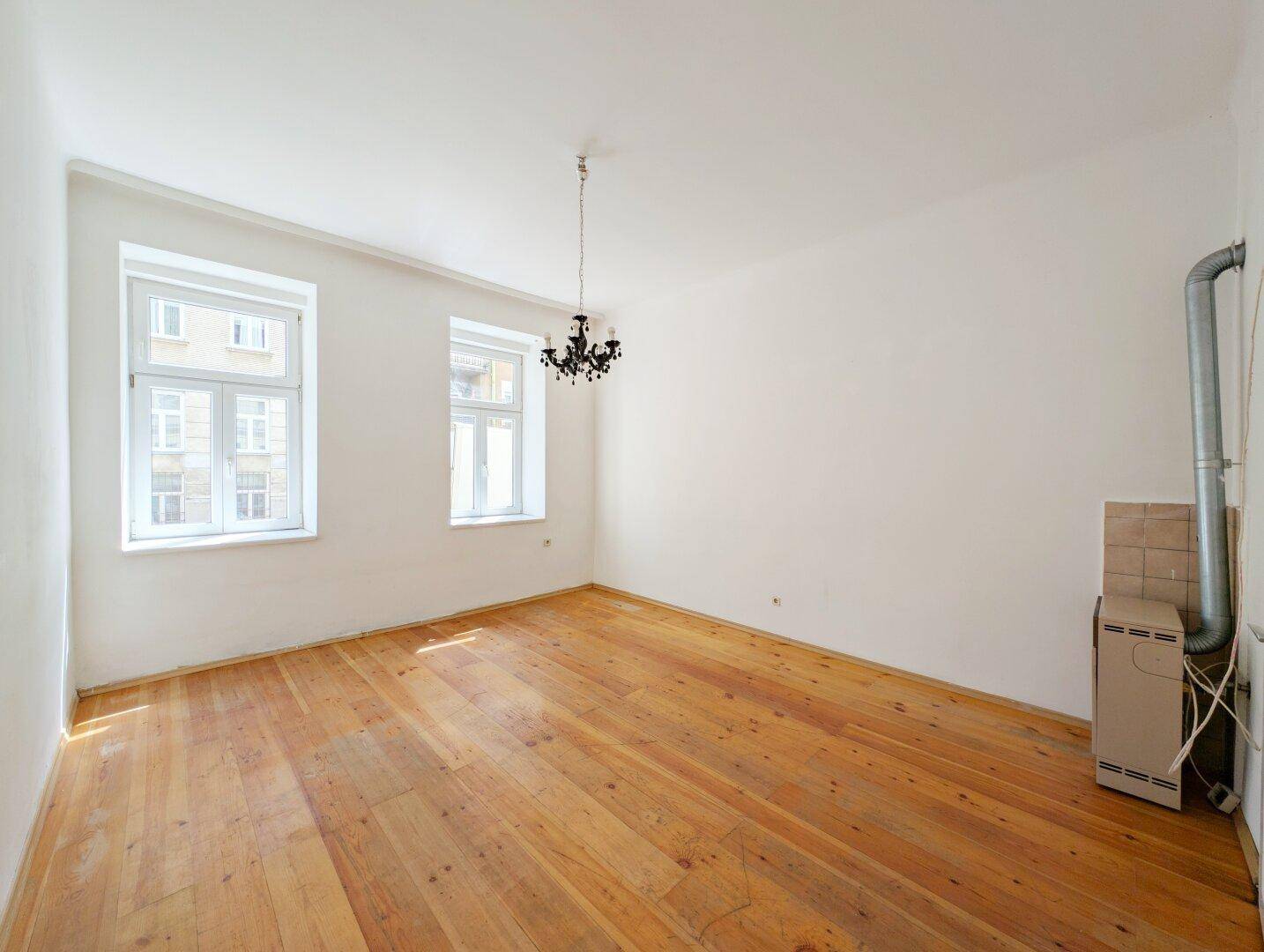 Geräumiges Zimmer mit Holzdielenboden, zwei Fenstern und einem sichtbaren Heizkörper.