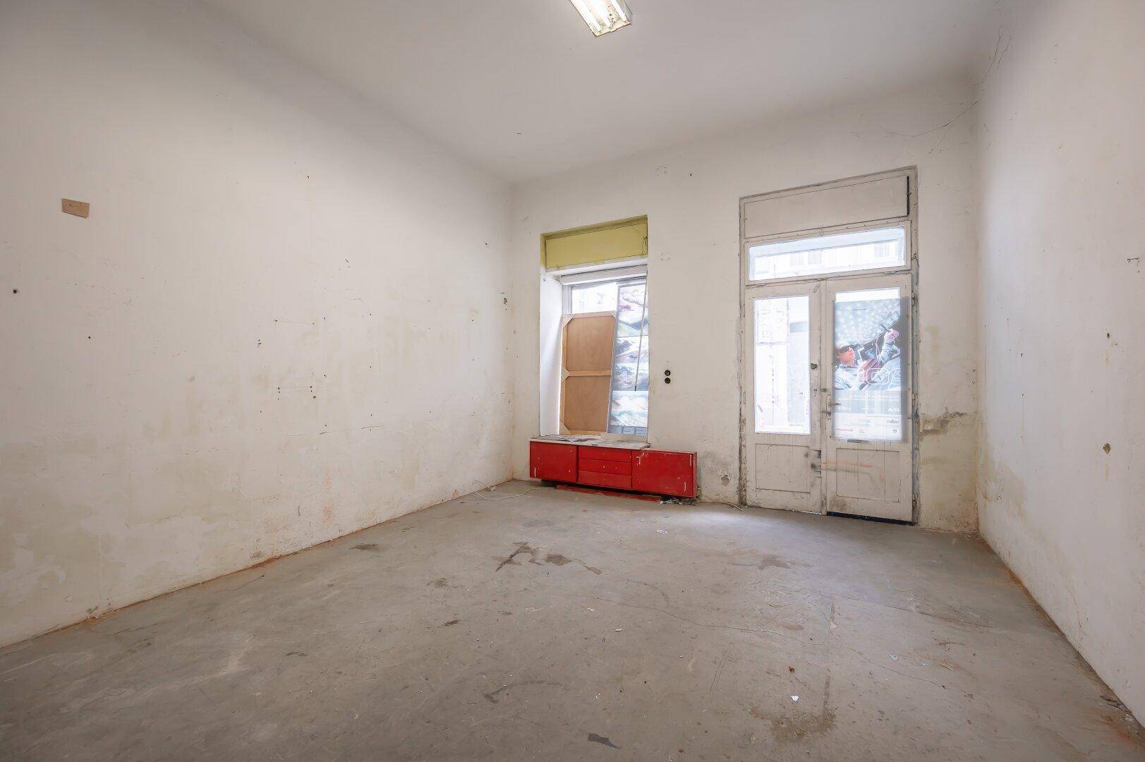 Leerer Raum mit abgenutztem Betonboden und einer alten Tür sowie einem Fenster zur Straße.