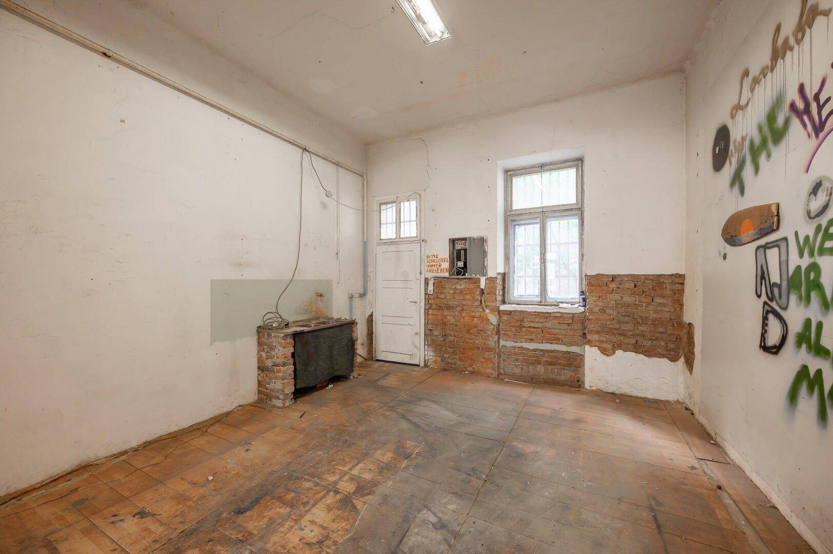 Leerer Raum mit abgenutztem Holzboden, Graffiti an der Wand und einem Fenster.
