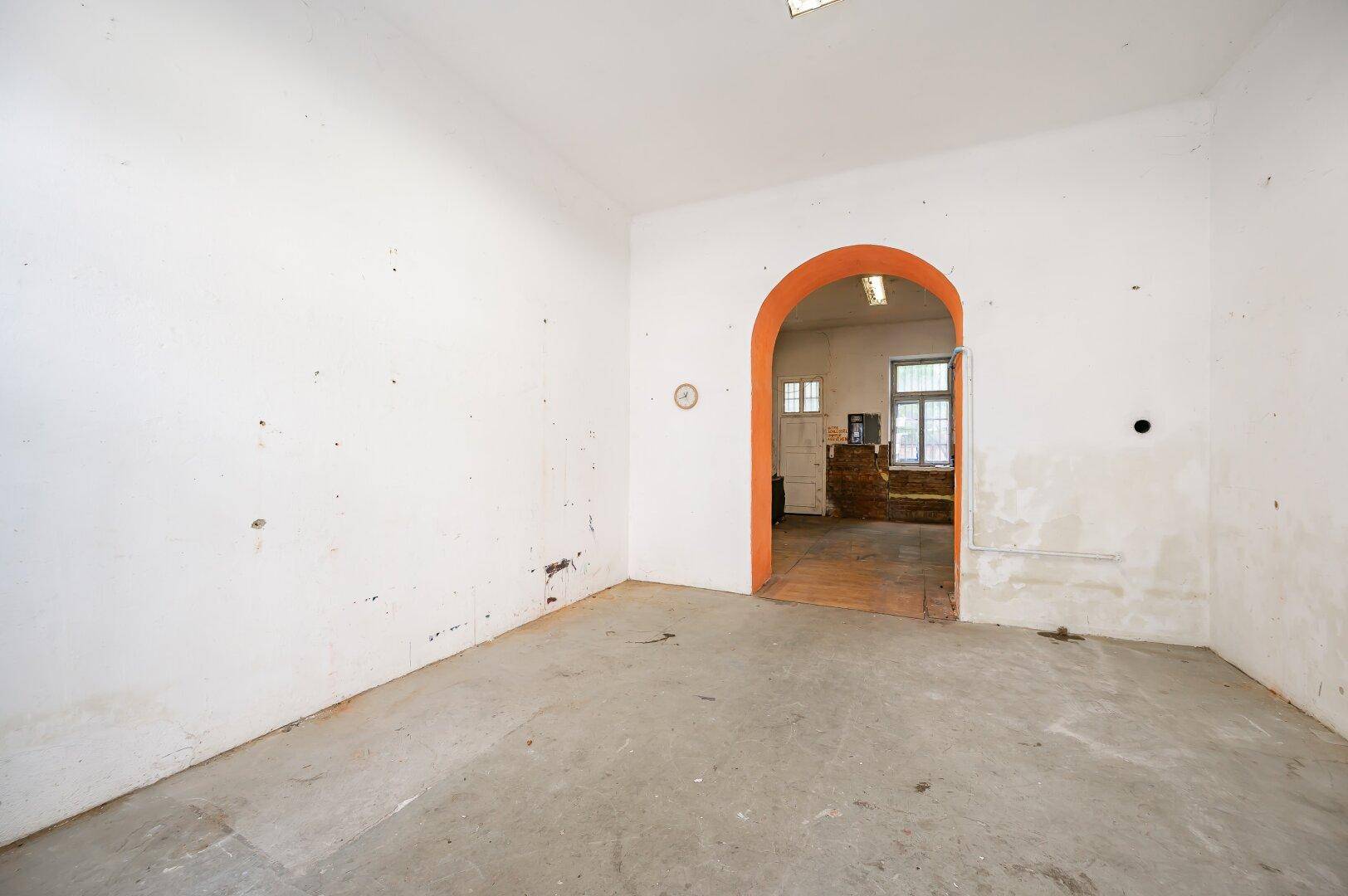 Leerer Raum mit Betonboden und einem orangefarbenen Torbogen, der zu einem weiteren Raum führt.