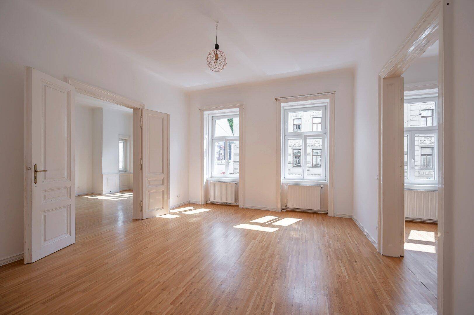 Geräumiges, leeres Zimmer mit Holzboden und viel Tageslicht durch große Fenster.
