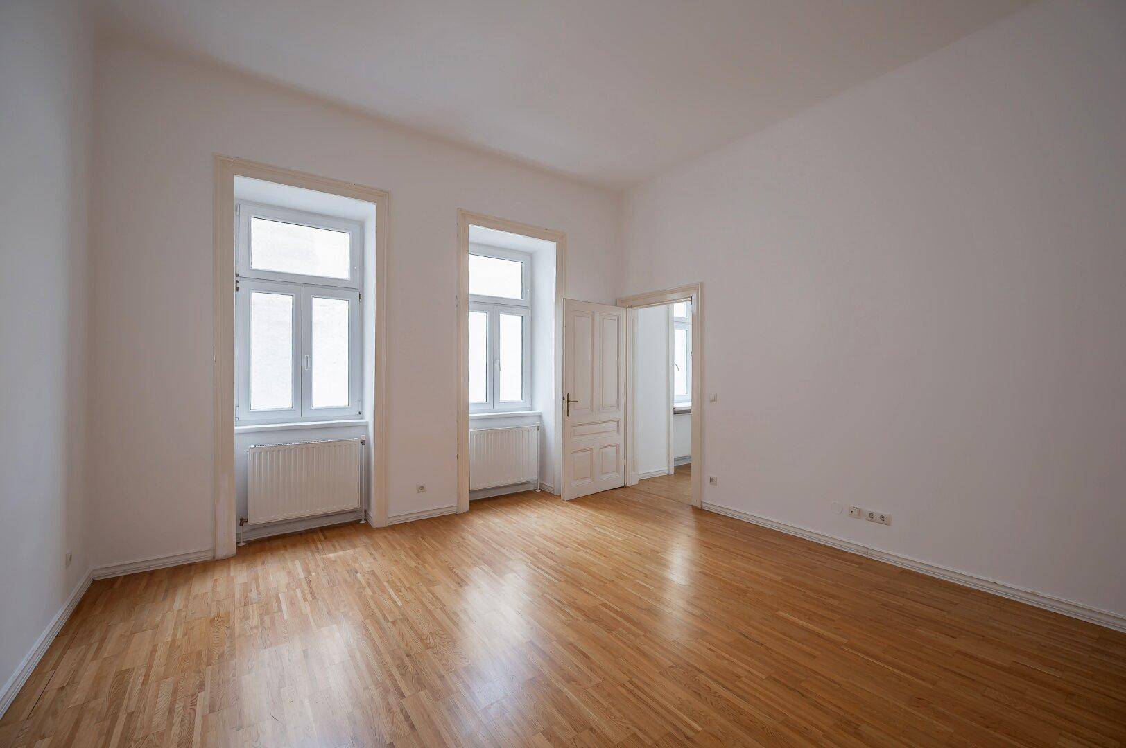Heller, leerer Raum mit Holzboden und zwei großen Fenstern, die Tageslicht spenden.