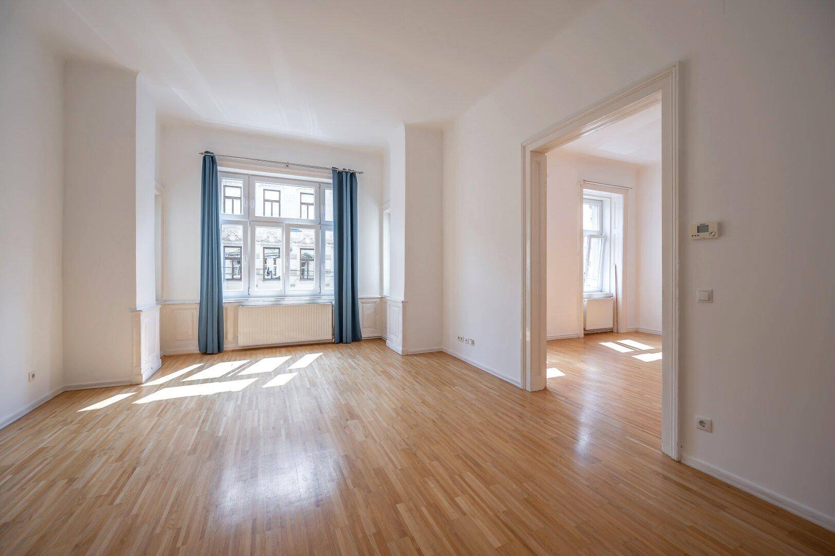 Helles, leeres Zimmer mit Holzboden, blauen Vorhängen und hohen Decken.