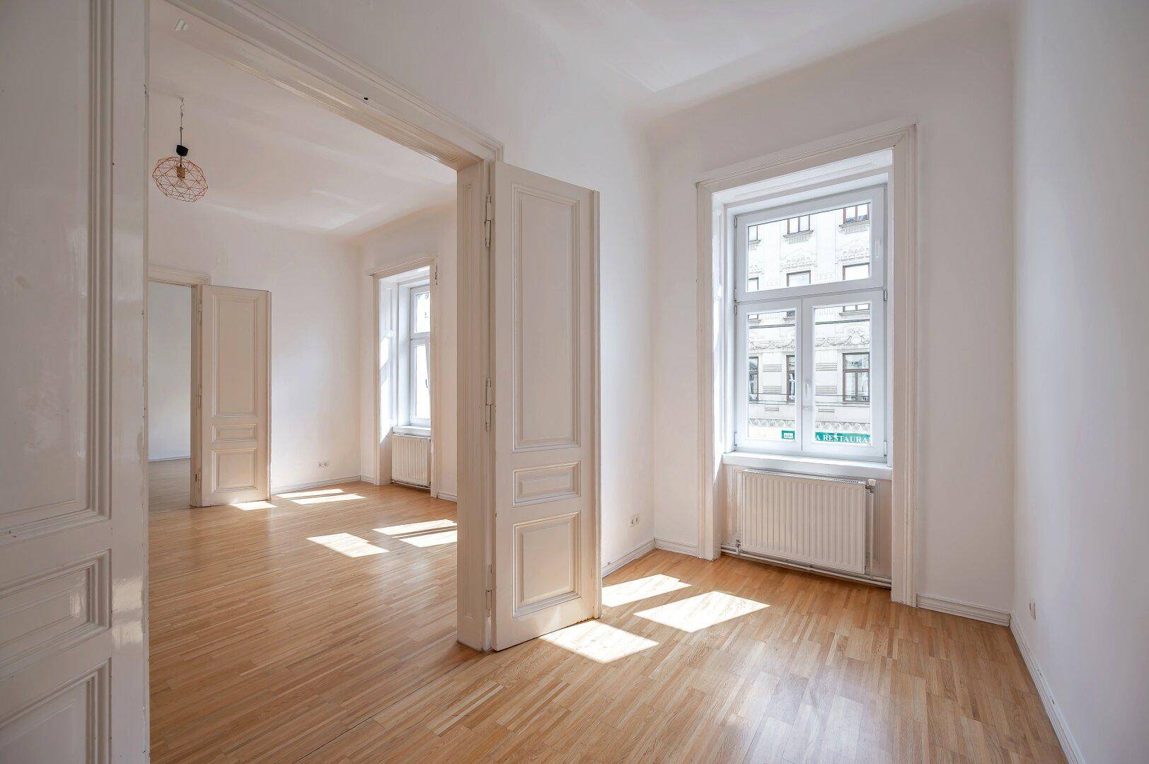 Leerer Raum mit Holzboden, hohen Türen und Fenstern, die viel Licht hereinlassen.