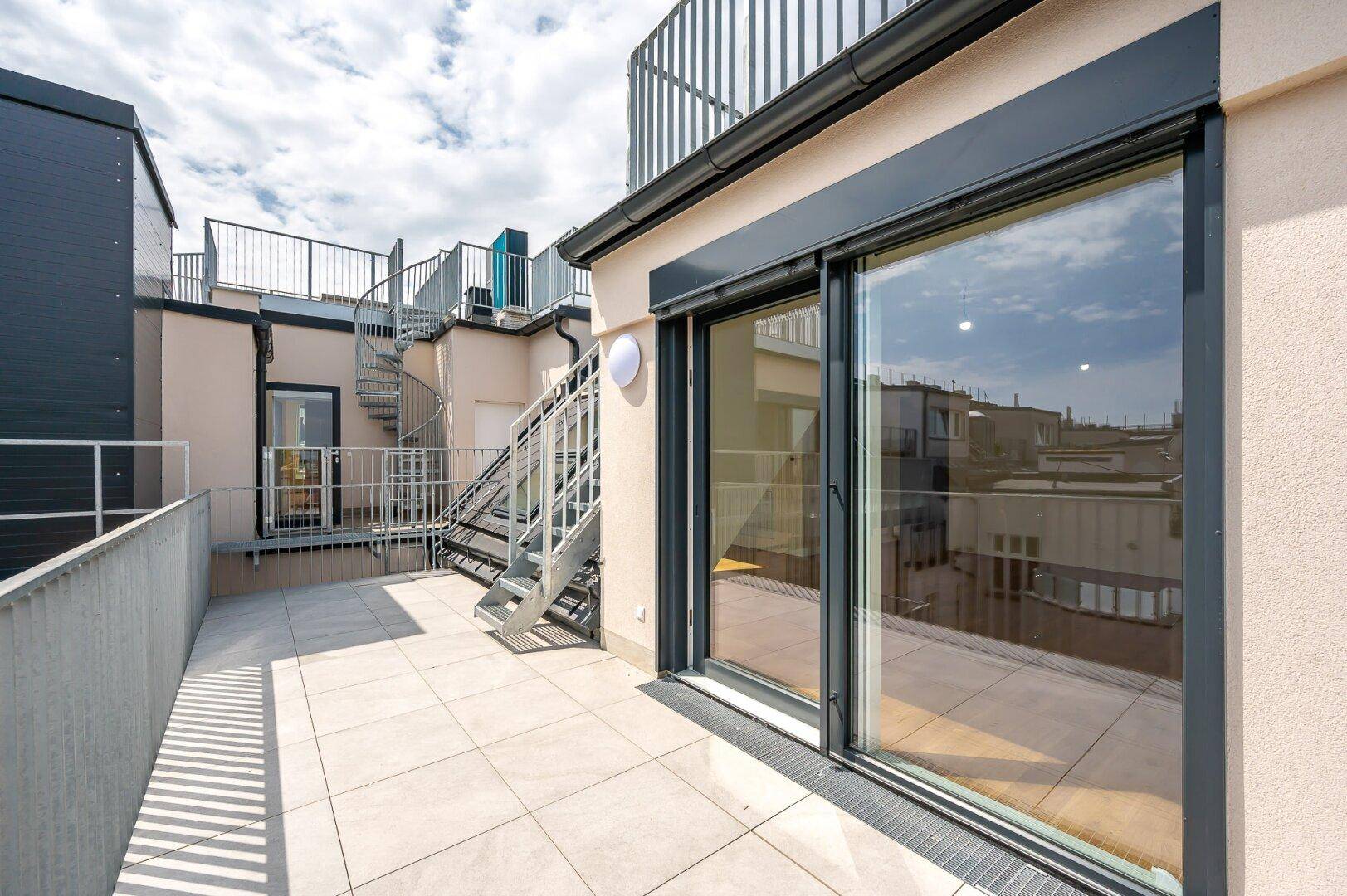 Heller Balkon mit Fliesenboden, Glasschiebetür und Treppe zur oberen Ebene.