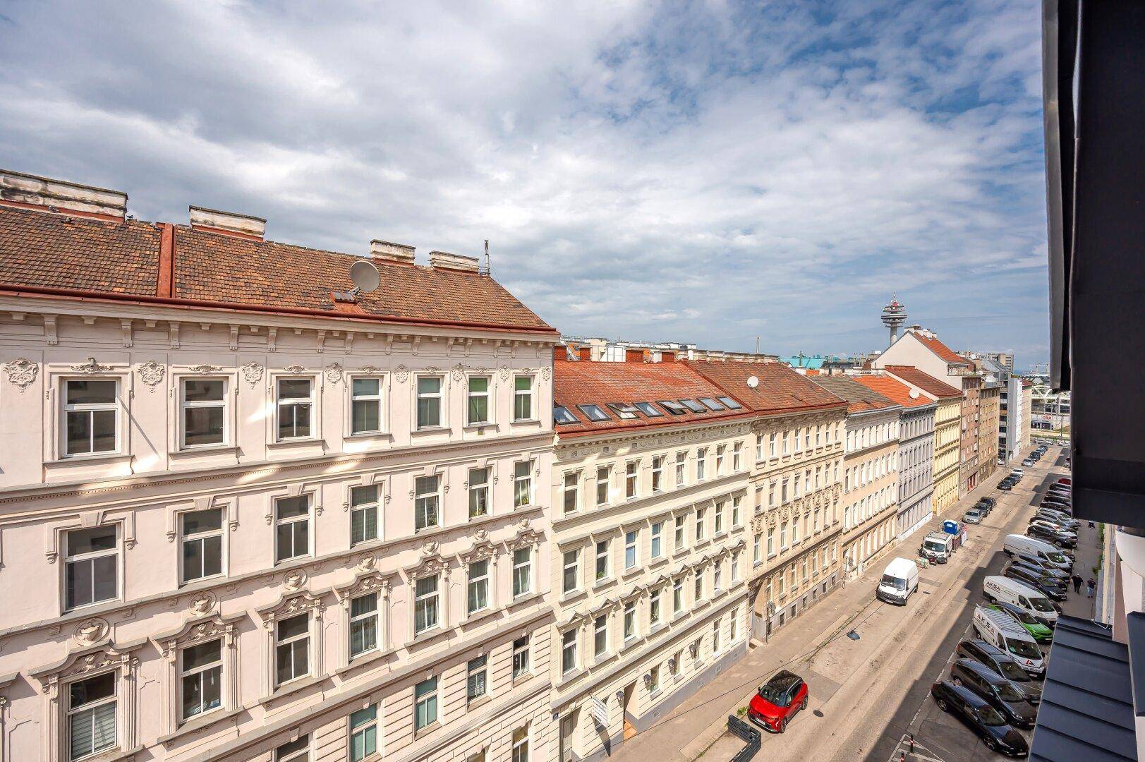 Panoramablick auf eine belebte Stadtstraße mit parkenden Autos und historischen Gebäudefassaden.