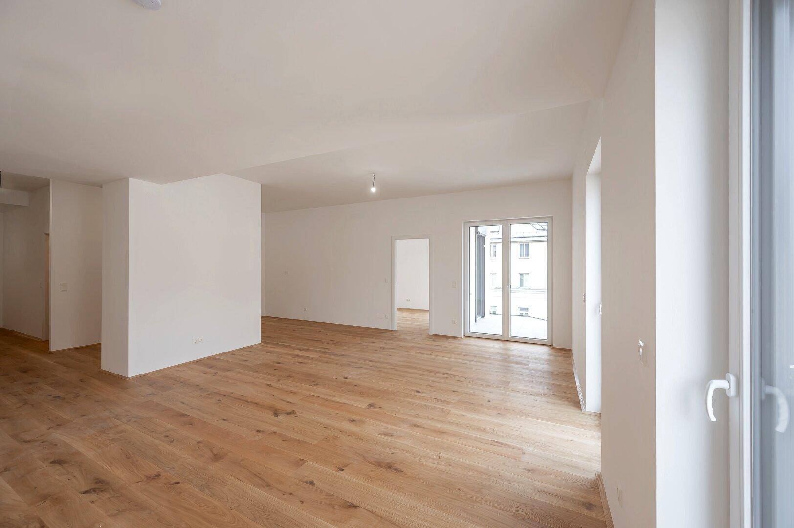 Heller, offener Wohnbereich mit Parkettboden und Zugang zu einem Balkon oder einer Terrasse durch eine Glasschiebetür.