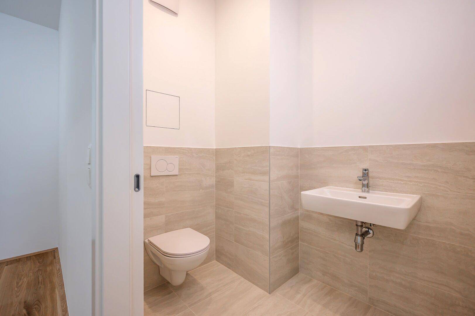 Separates Gäste-WC mit Wand-WC, kleinem Waschbecken und beigefarbenen Fliesen.