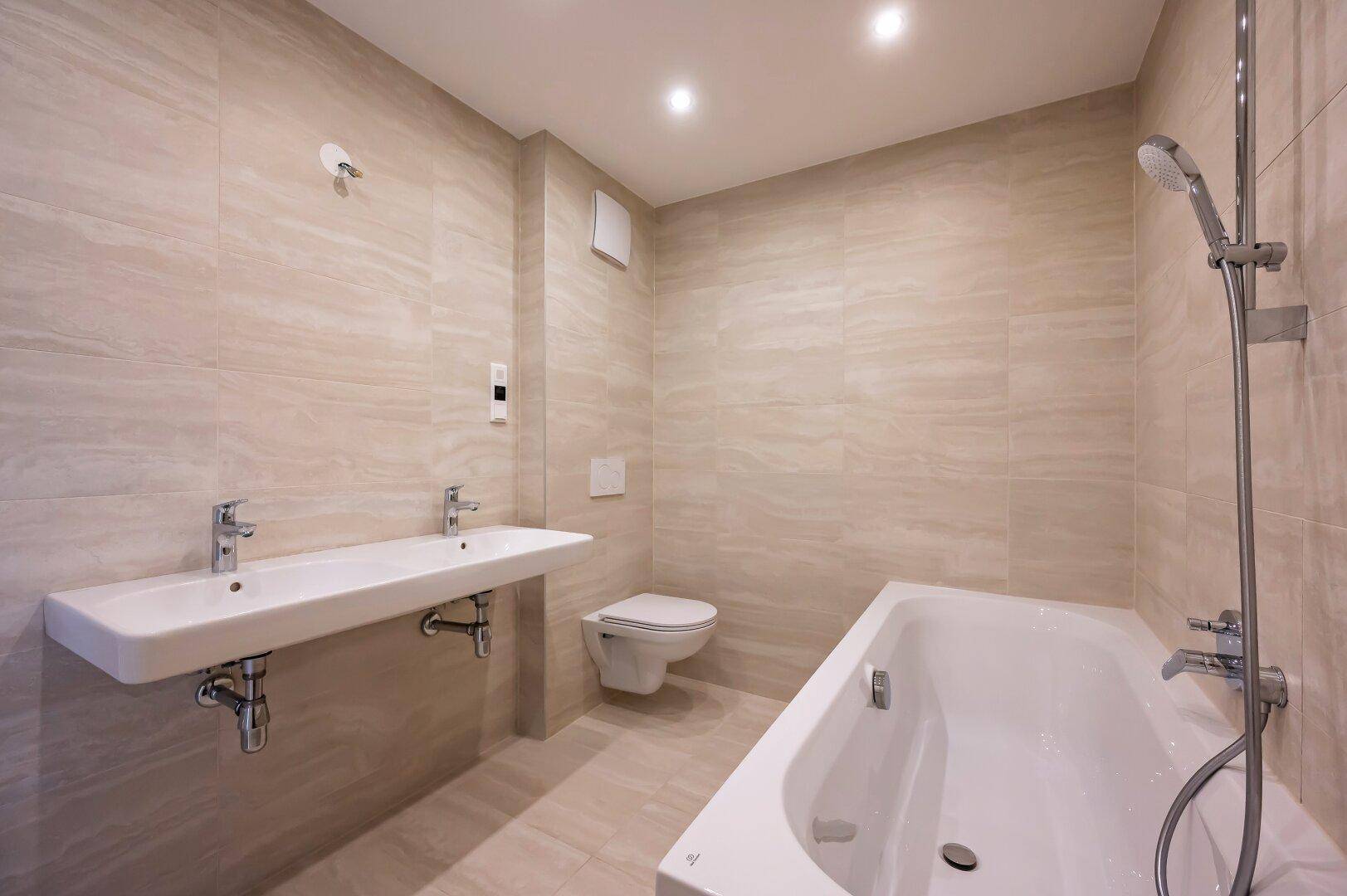 Geräumiges Badezimmer mit Doppelwaschbecken, Badewanne und hellen, modernen Fliesen an den Wänden.