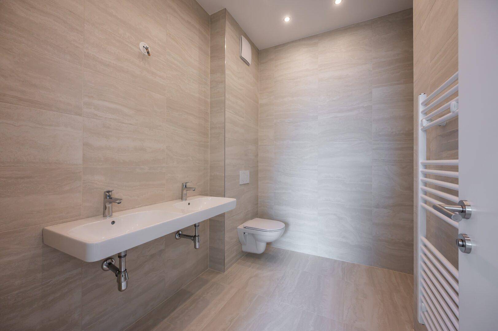 Modernes Badezimmer mit doppelten Waschbecken, Wand-WC und beigen Fliesen.